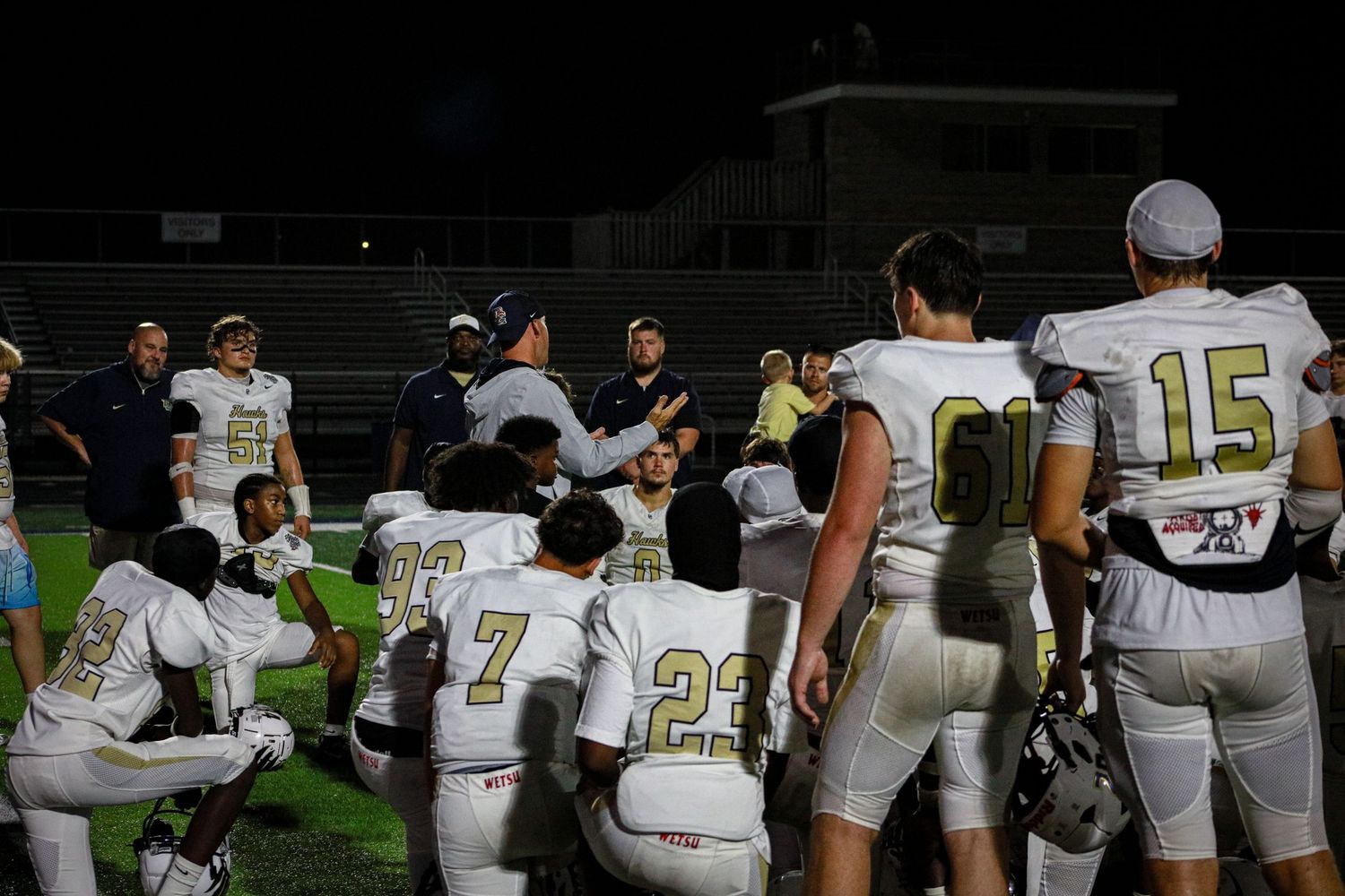 High school football players in white uniforms huddle with coaches on the field during a night game.