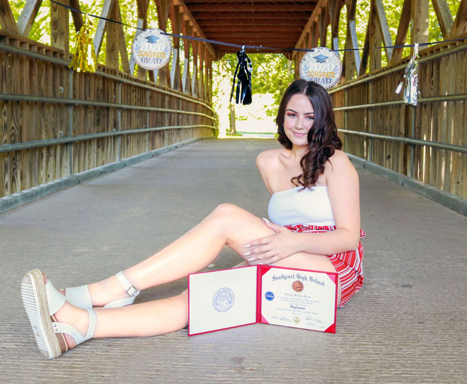 Young woman in graduation attire poses on a wooden covered bridge with her high school diploma and awards.
