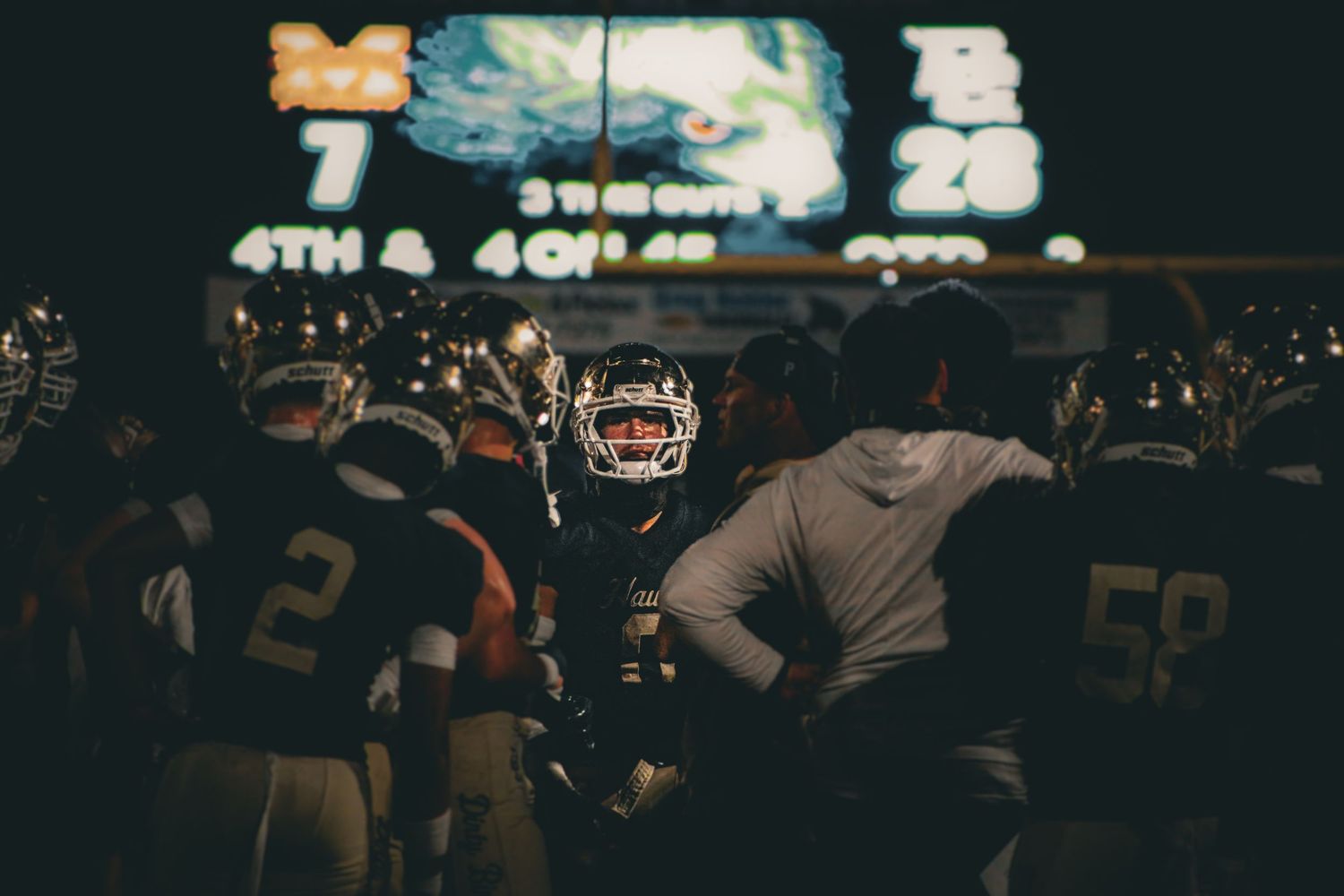 Football players huddle near tunnel under scoreboard showing 4th down during a night game.