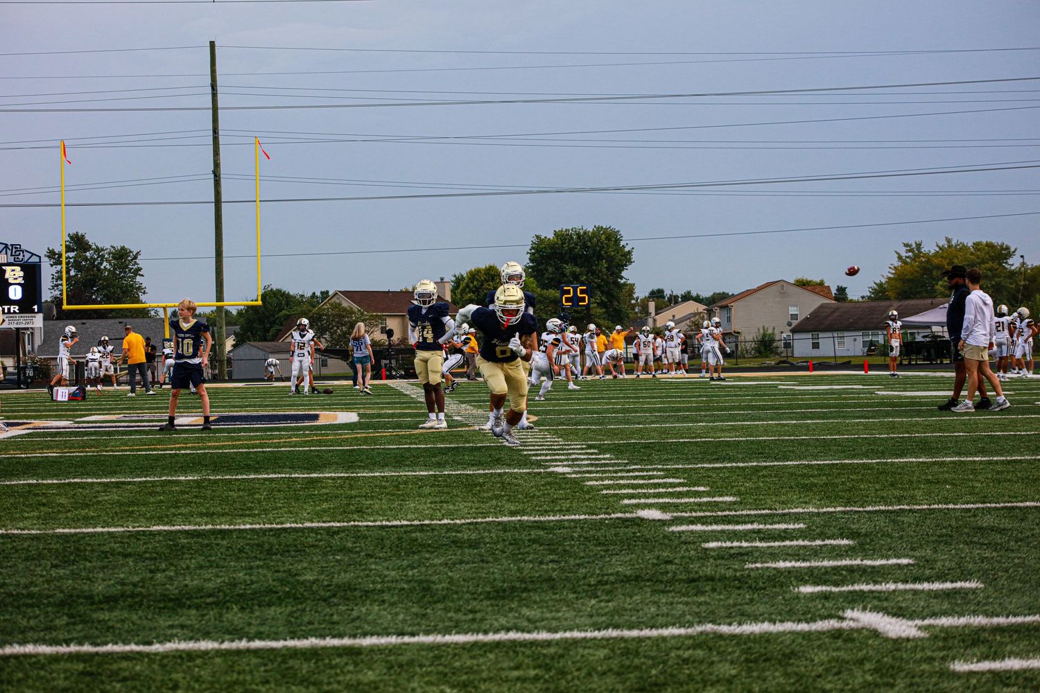 High school football players in gold and white uniforms practice drills on a green turf field during a sunny day.