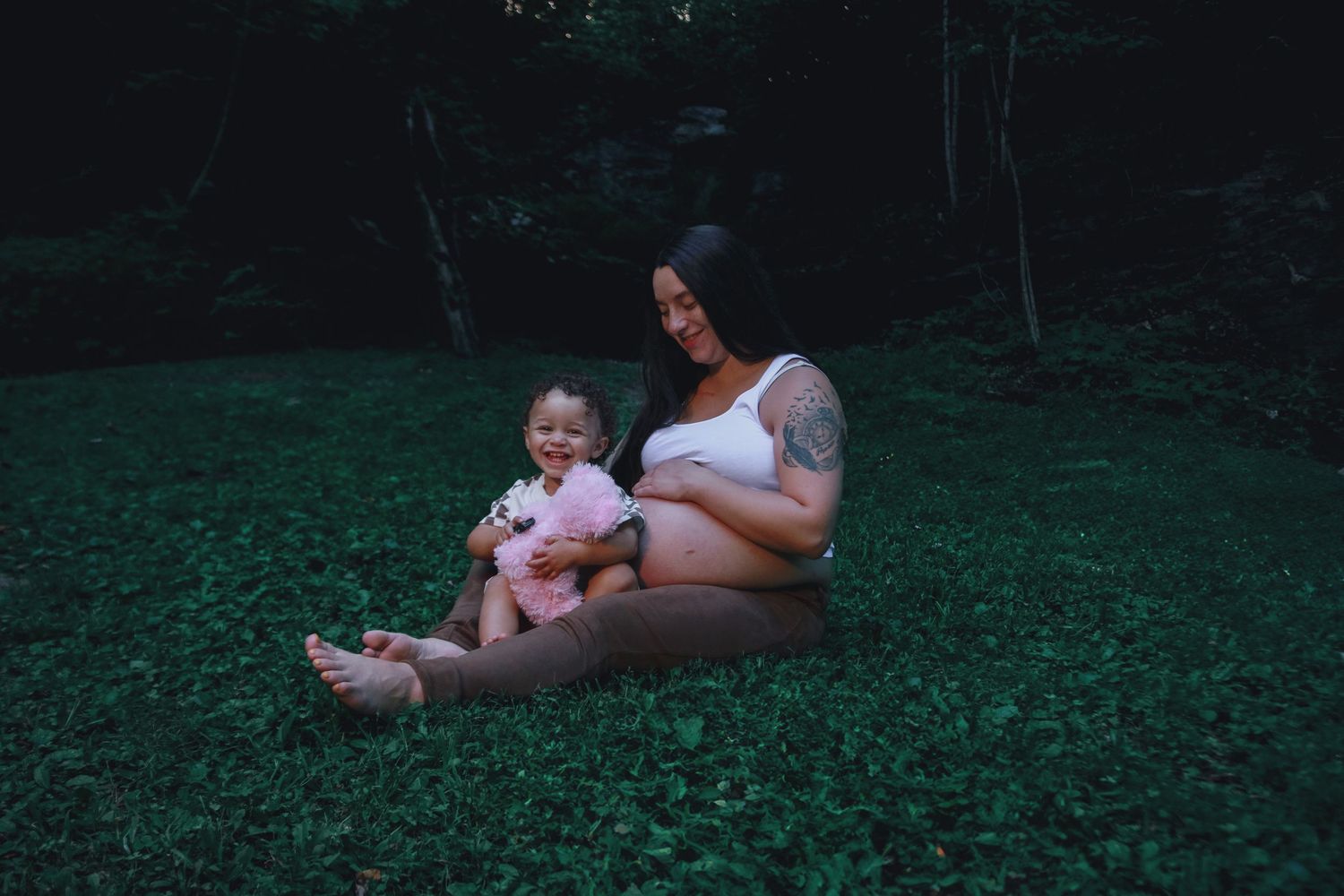 Pregnant mom holding pink teddy bear with son sitting on her lap.