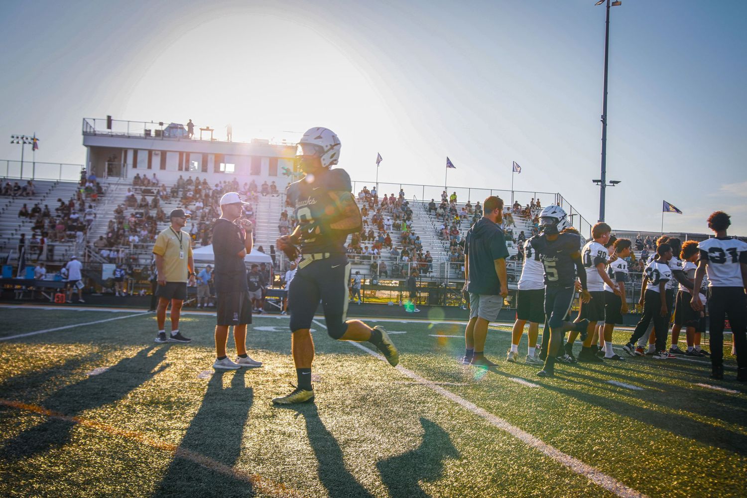 Football players warm up on a sunlit field during a high school game, with stadium bleachers and fans visible in background.