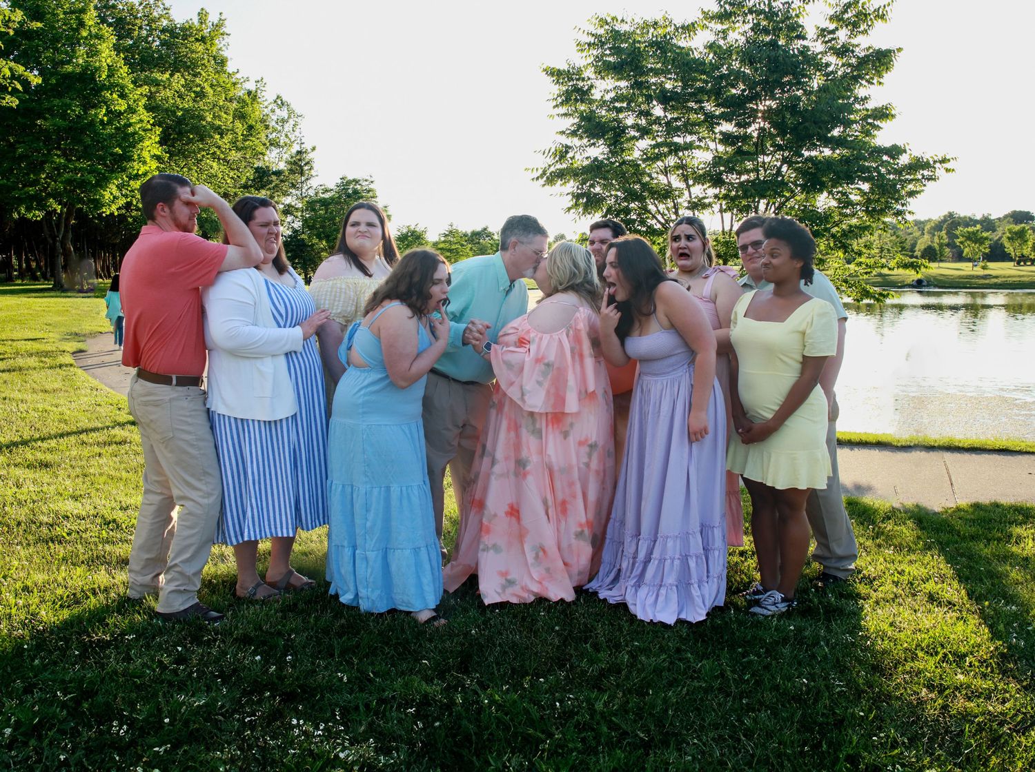 Group of people in pastel dresses gathering around kissing couple in sunny park.
