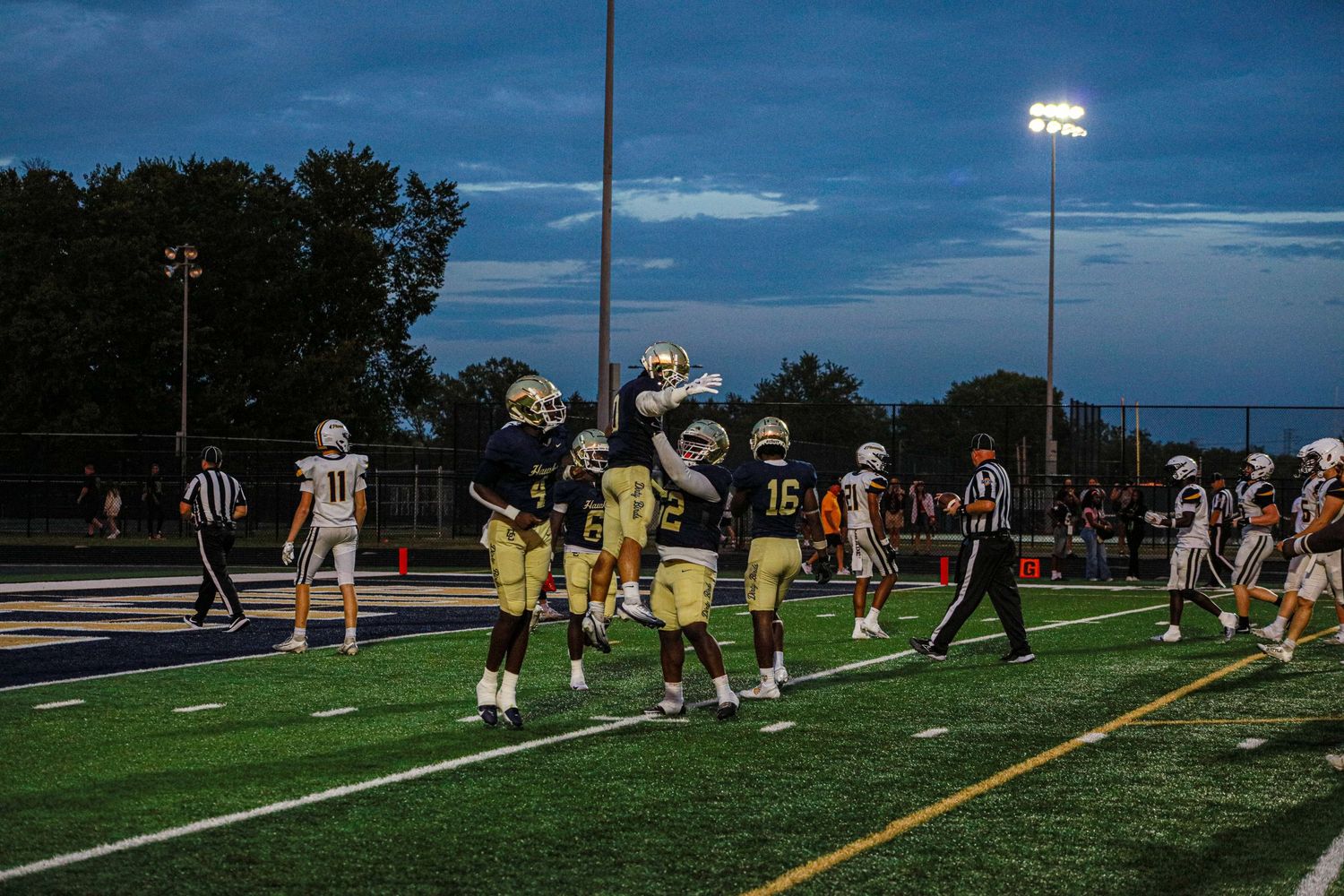 Football players in gold uniforms celebrate on a lit field during an evening high school football game.