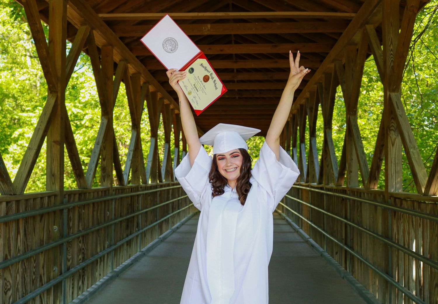 Graduate in white cap and gown celebrates on a covered wooden bridge, holding diploma overhead with arms raised.