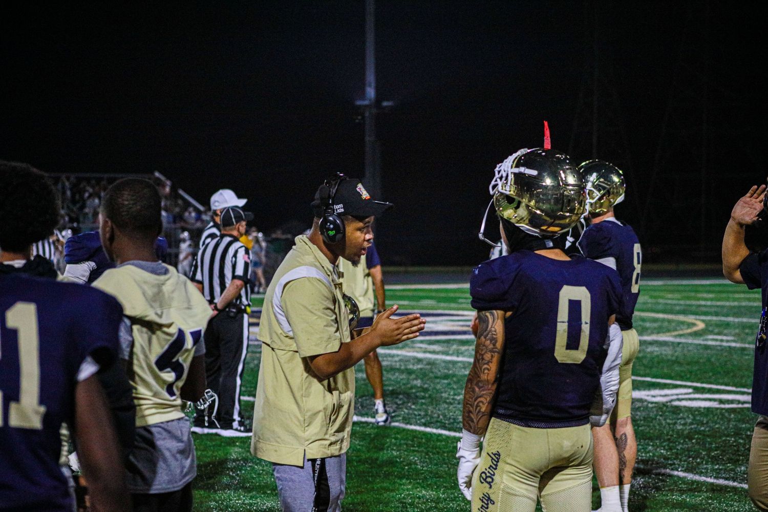 Football coach in headset talks to player #0 on sideline during night game under bright field lights.