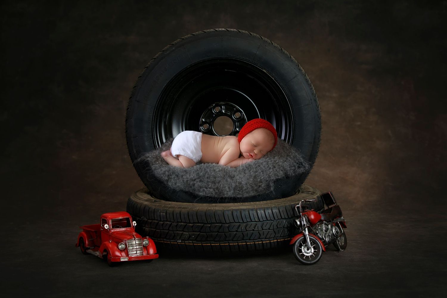 Newborn baby sleeping peacefully inside a tire prop surrounded by miniature toy vehicles.
