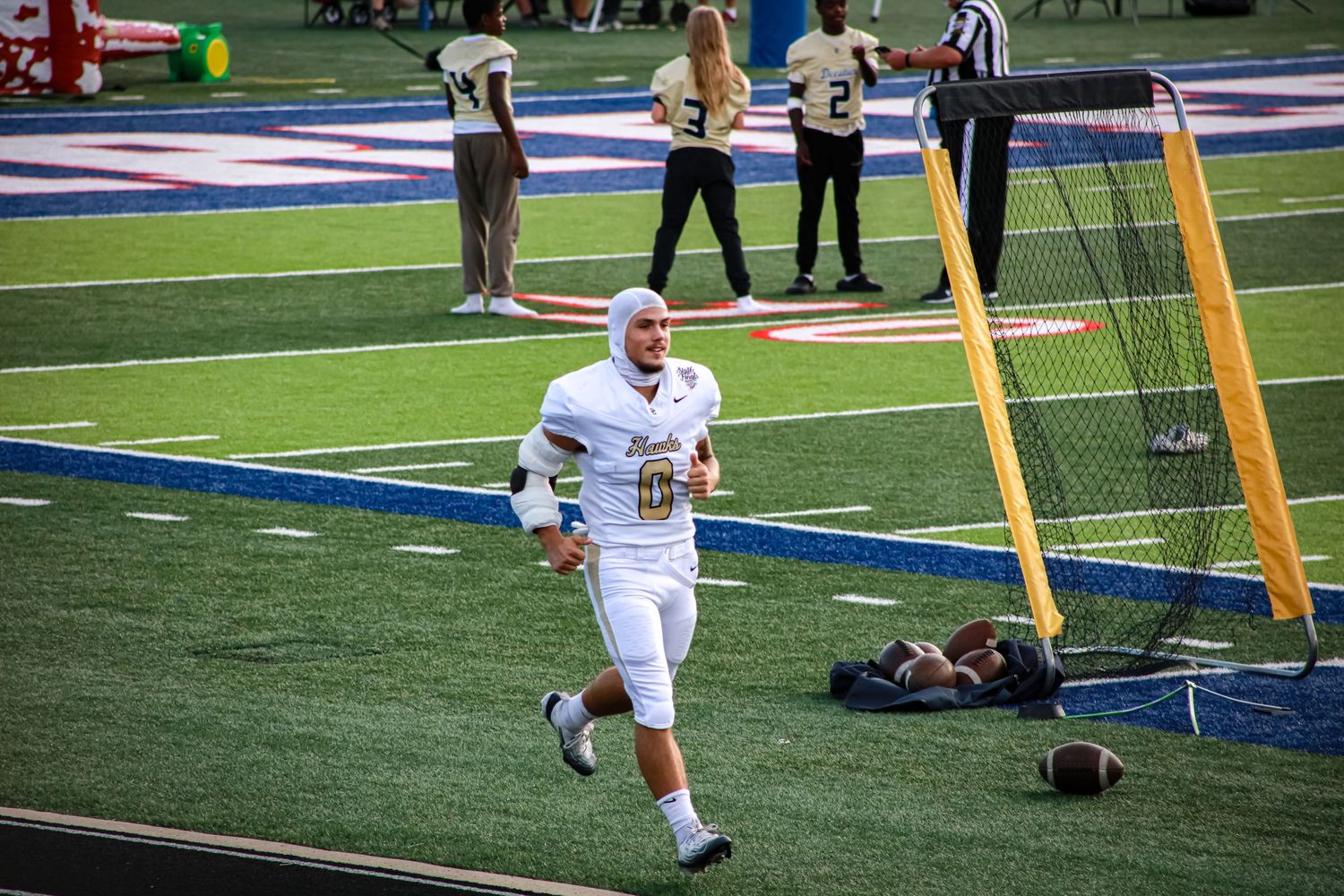 Football player in white uniform jogging on blue turf field during a game with goal post and referee in background.