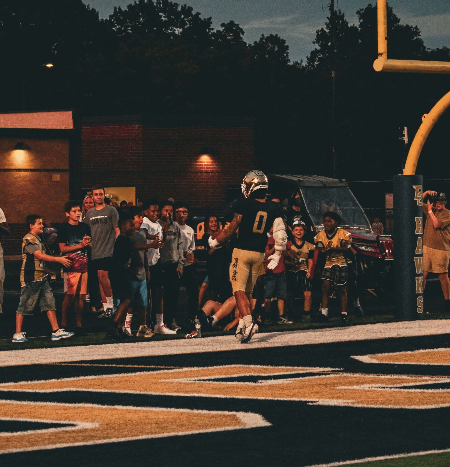 Football player wearing number 0 runs along sideline past cheering fans during a night game at a high school stadium.