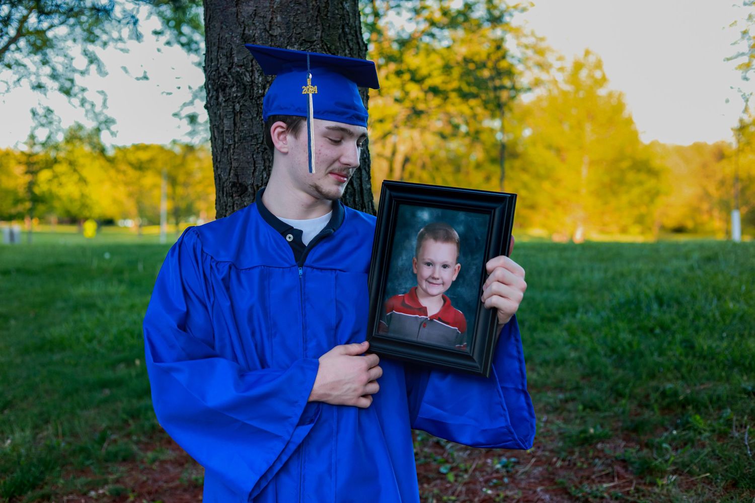 A young man in blue graduation cap and gown holds a framed childhood photo beside a tree.