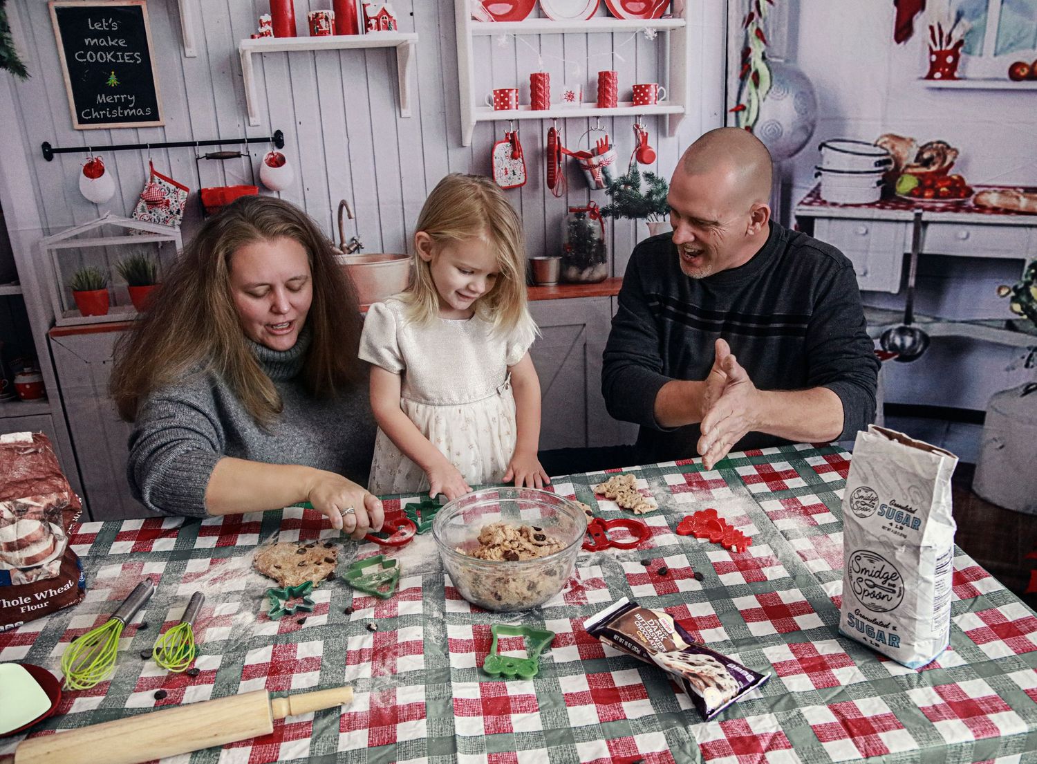 Family of three making Christmas cookies together at a festive holiday decorated kitchen table.
