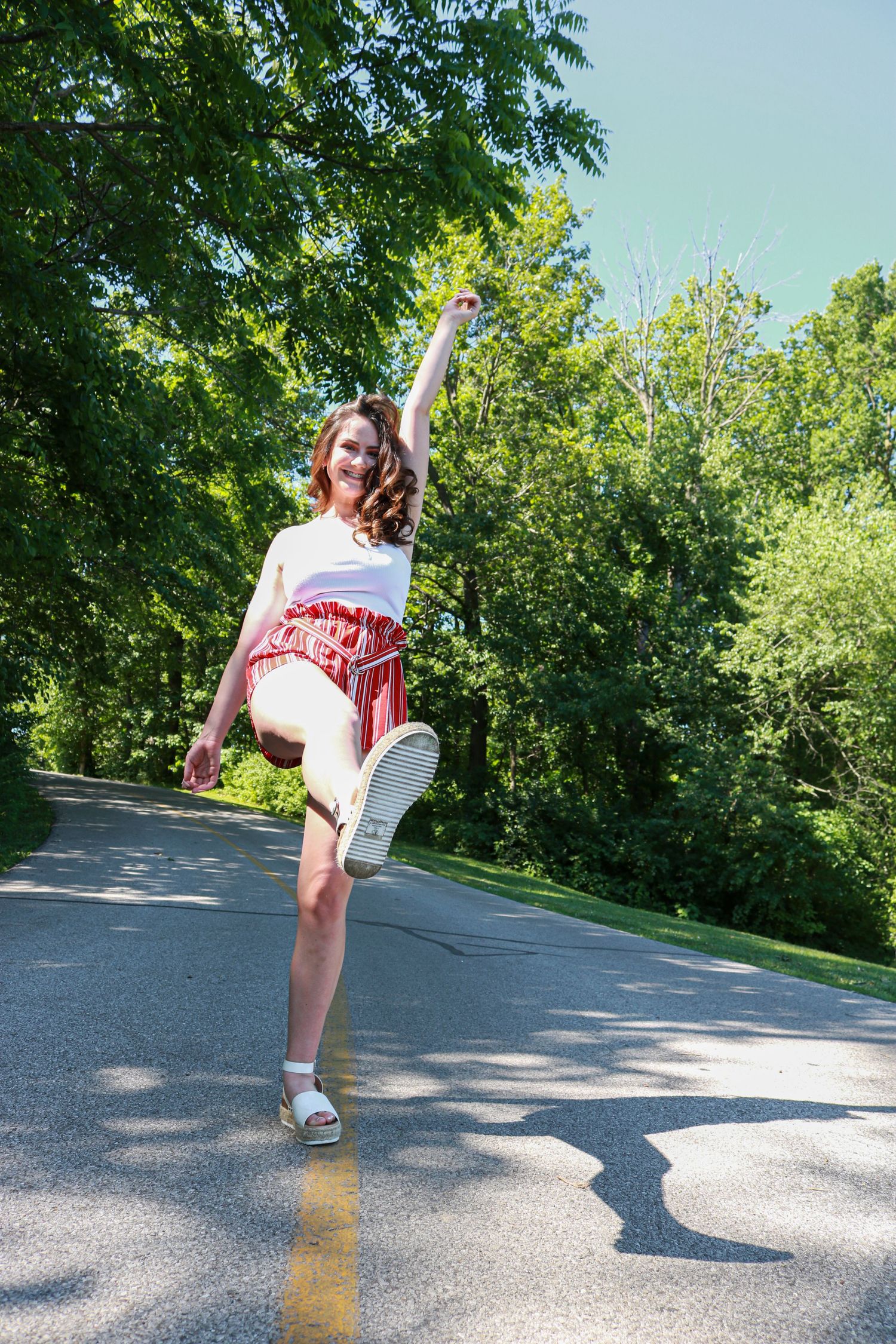 Young woman in red skirt and white top dancing joyfully on a tree-lined road on a sunny day.