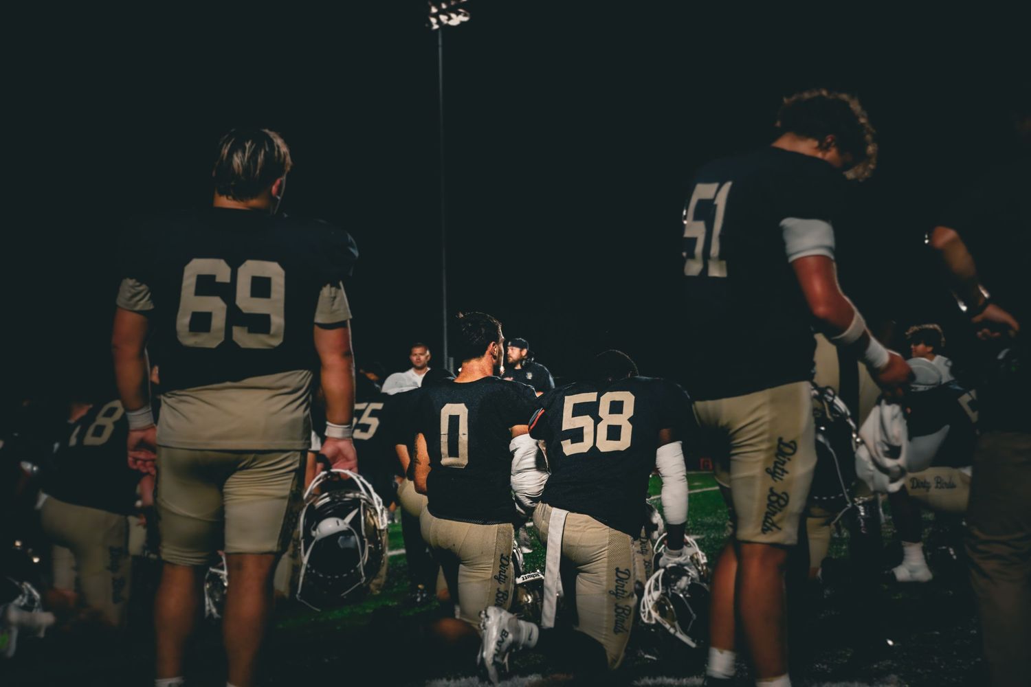 Football players in dark jerseys numbered 69, 0, 58, and 51 huddle on the sideline during a night game.