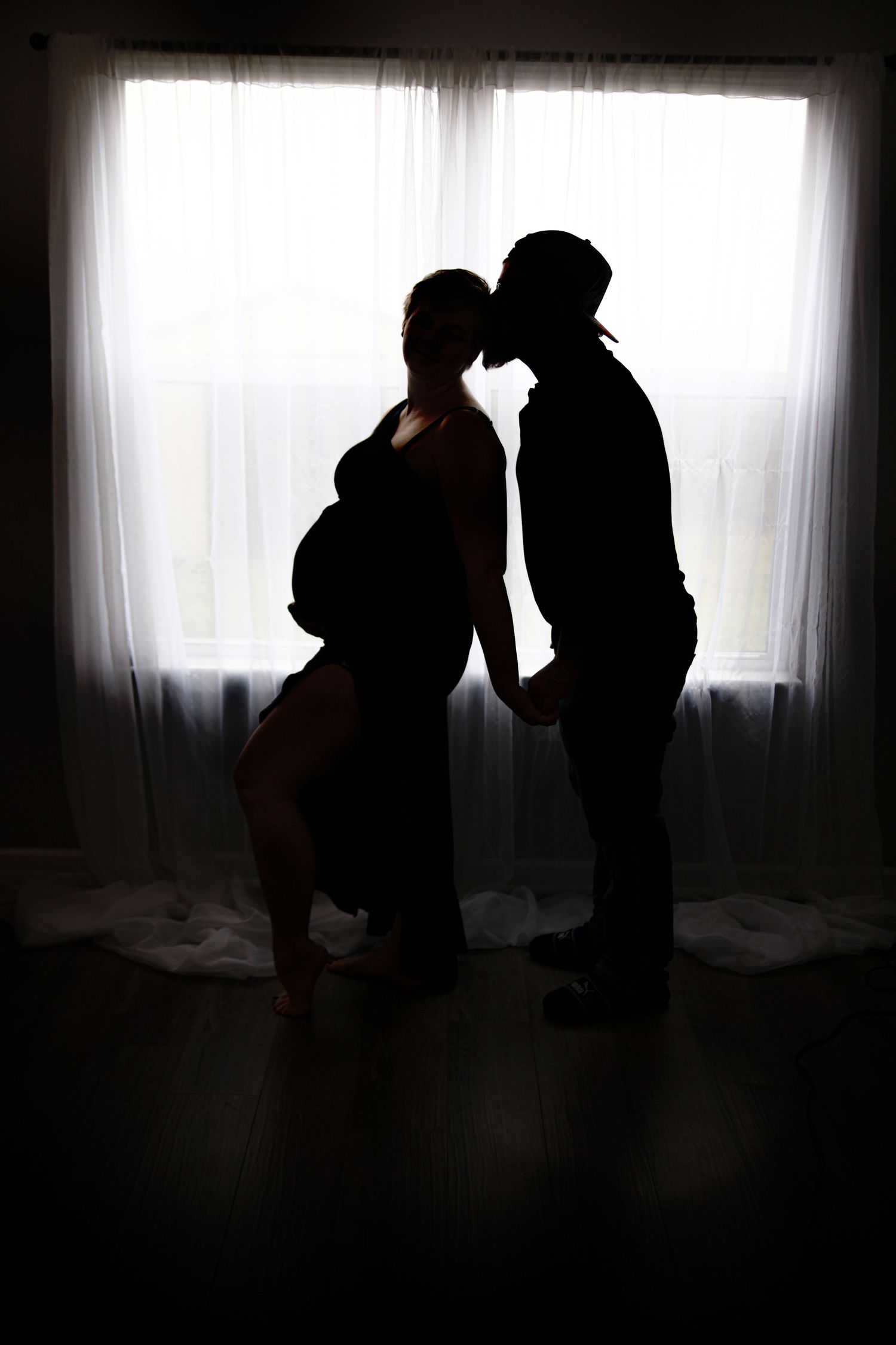 Silhouette of pregnant couple kissing in front of bright window with sheer curtains.