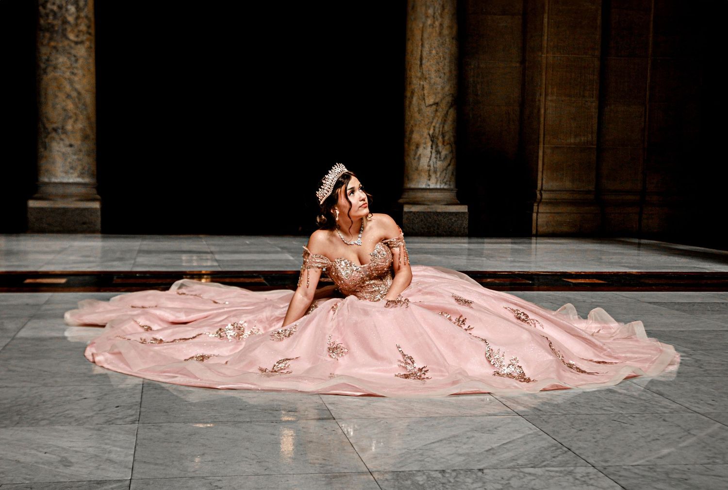 Young woman in elegant pink ballgown with floral embroidery poses gracefully on marble floor in grand hall.