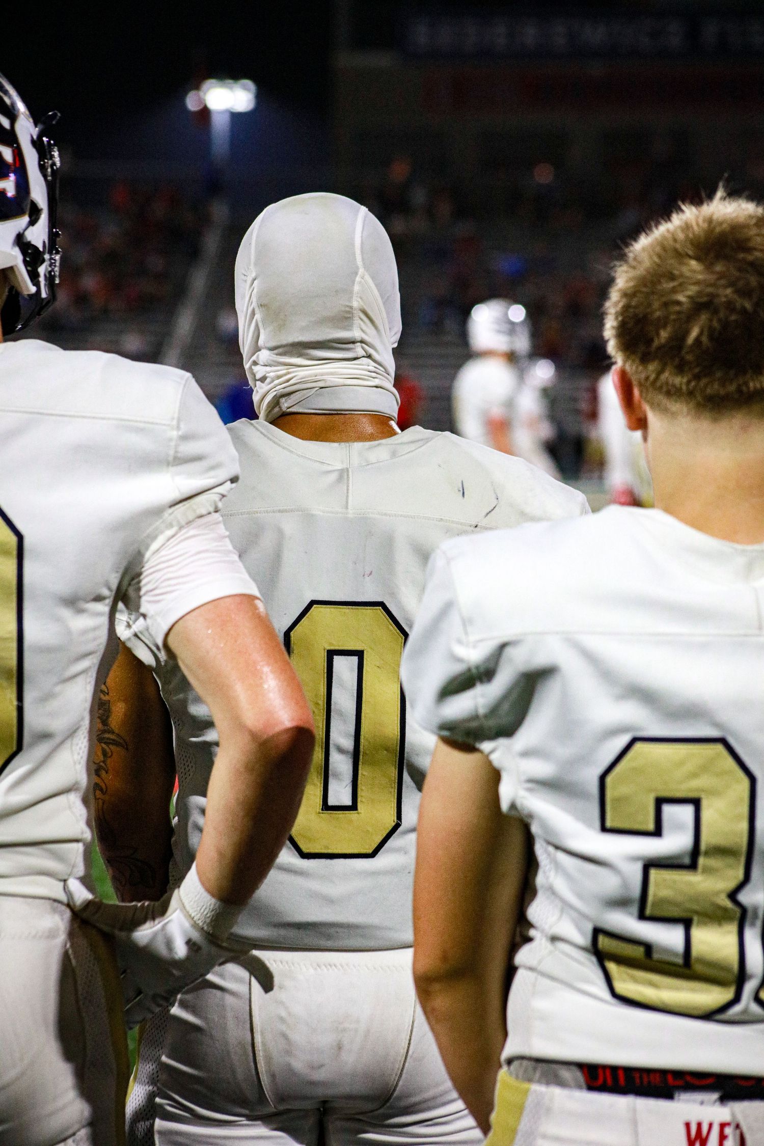 Football players in white and gold uniforms stand on the sideline during a night game.
