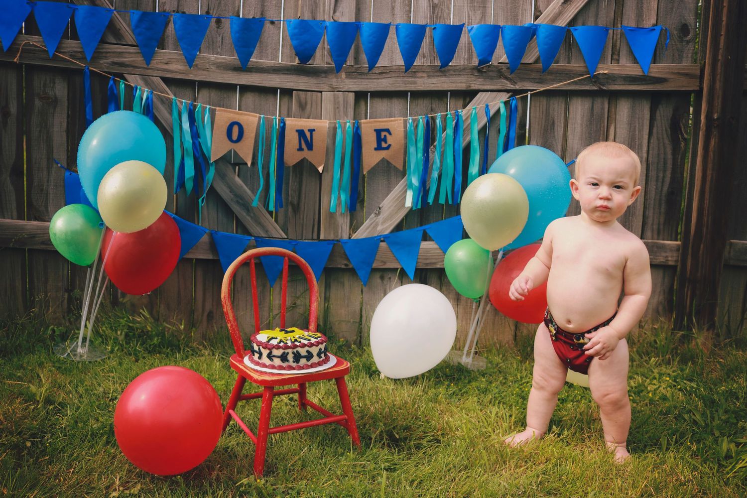 One year old boy standing outside with birthday decorations and birthday cake.