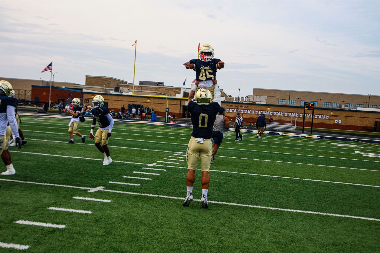 Football players in gold and black uniforms warm up on a green turf field during a high school game.