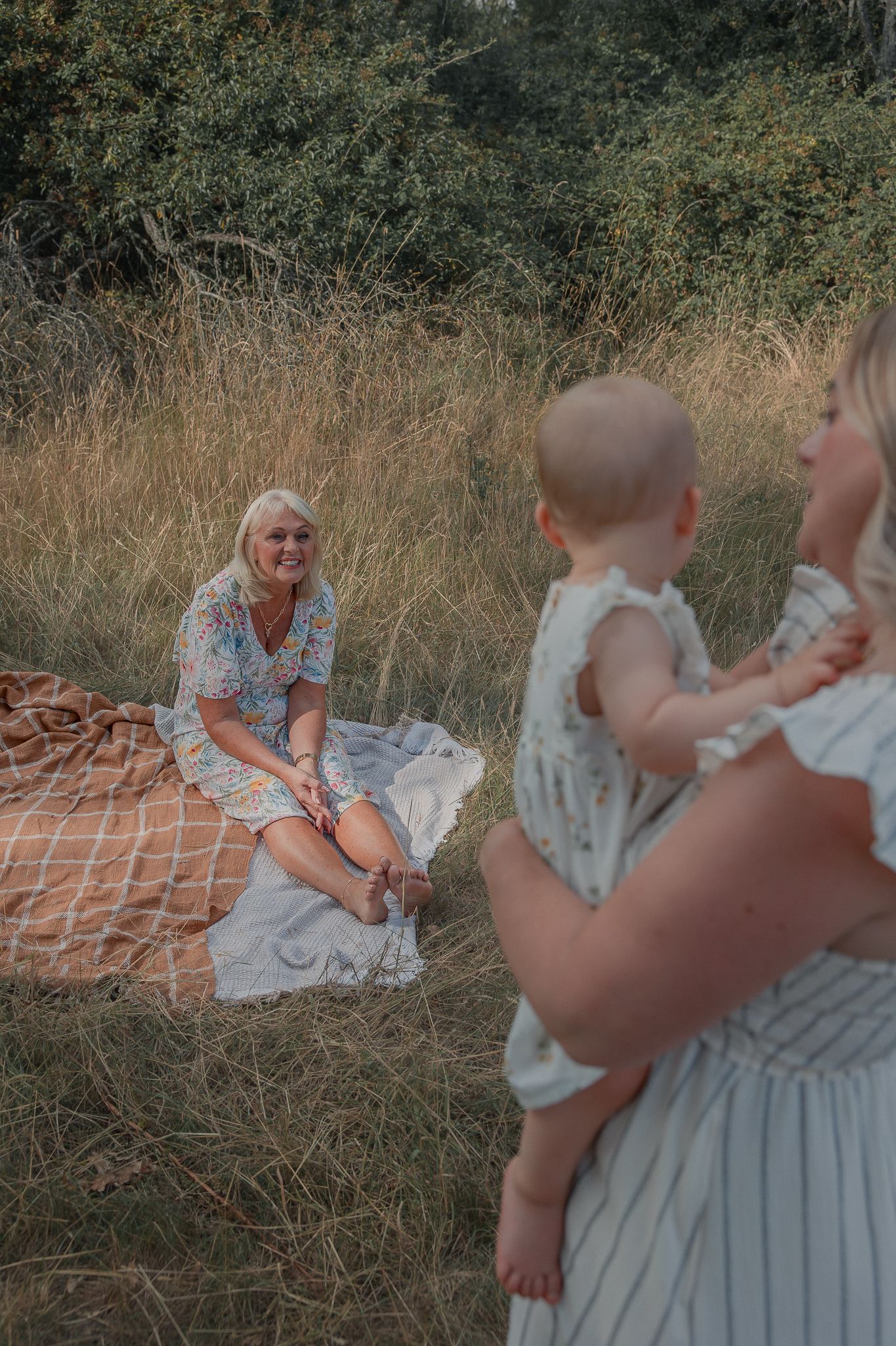 Smiling blonde woman sitting on blanket in field with mother holding baby.