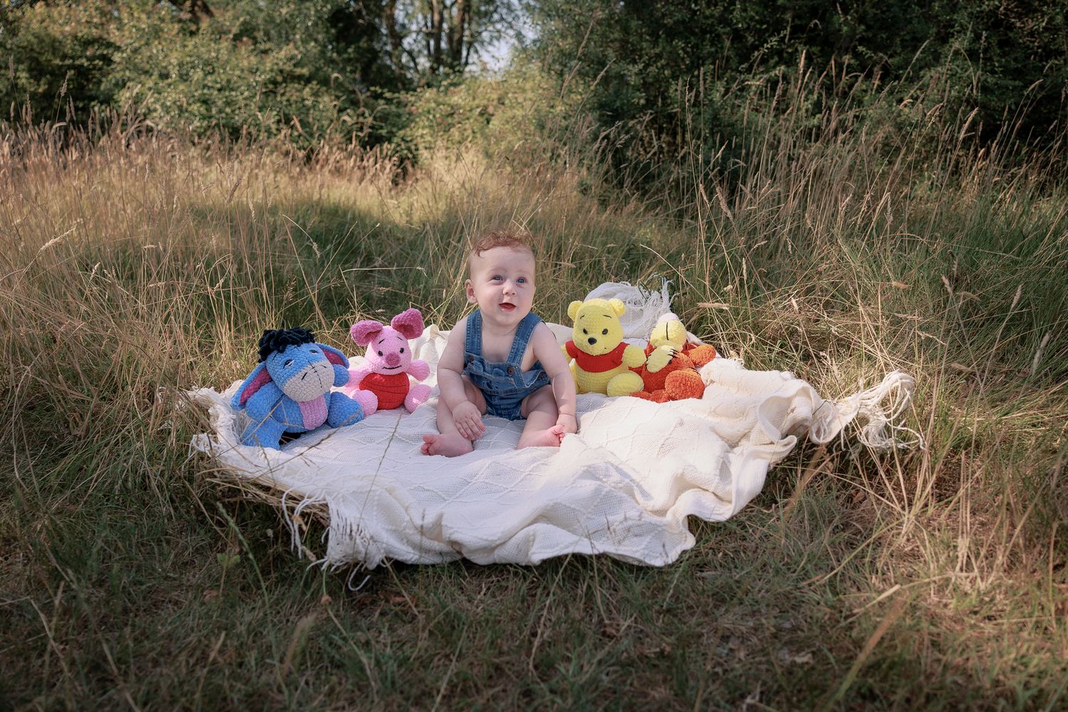 Baby sitting on white blanket in grass field surrounded by Winnie the Pooh stuffed animals toys outdoors in Stow, Cotswolds.
