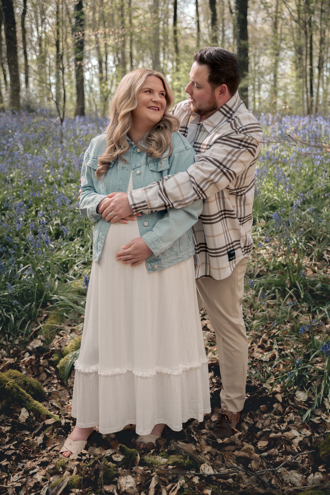 Couple embracing in a bluebell woodland, pregnant woman in white dress and denim jacket with partner in plaid shirt.