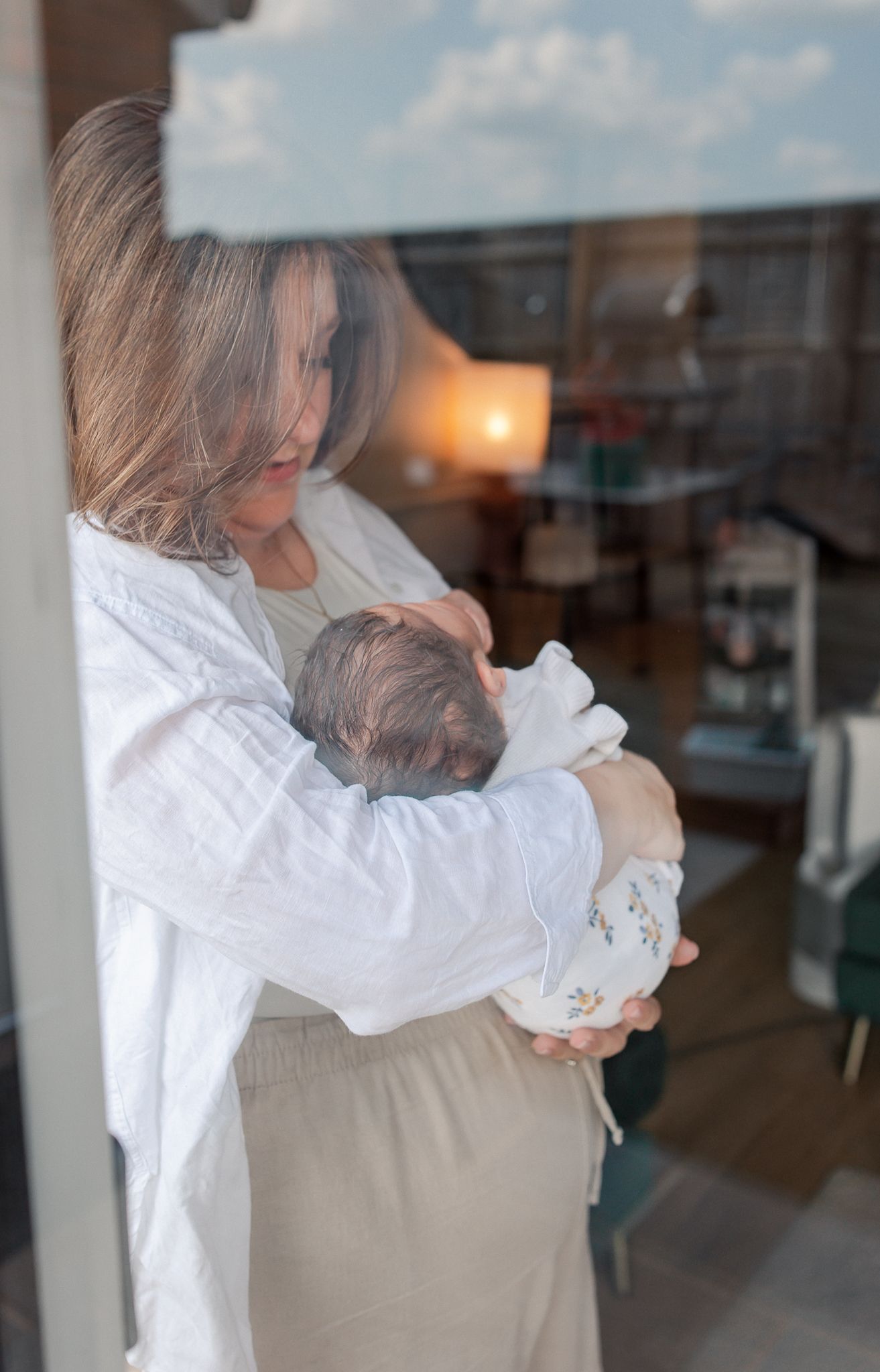 A mother tenderly holds her newborn baby wrapped in white near a window with soft indoor lighting in Bradford-on-Avon.