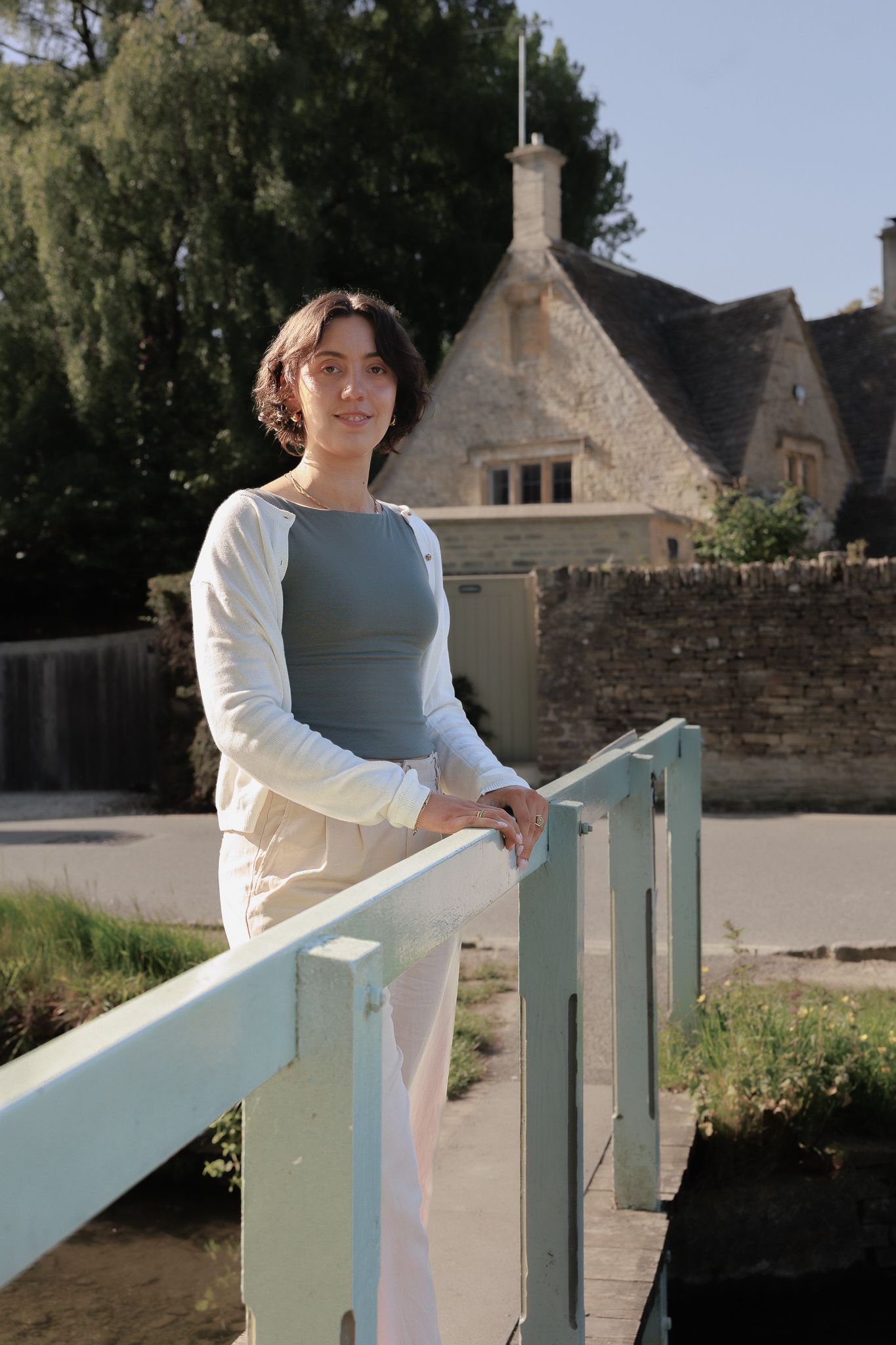 Young woman leaning on a light green bridge railing near a stone cottage in Lower Slaughter near Stow-on-the-wold.