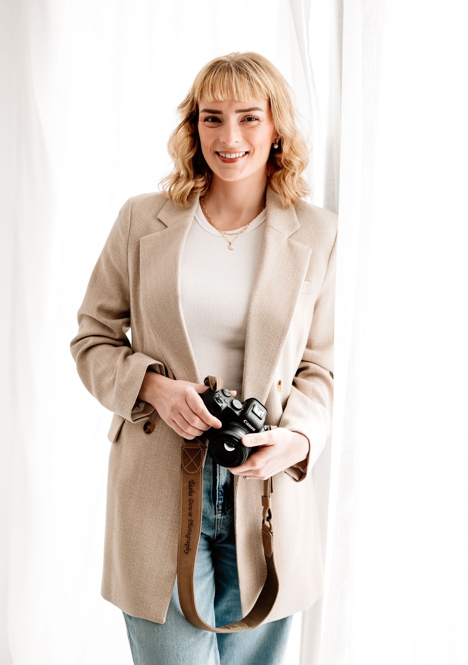 Smiling blonde woman in beige blazer and jeans holding a camera with strap, standing near white curtains.