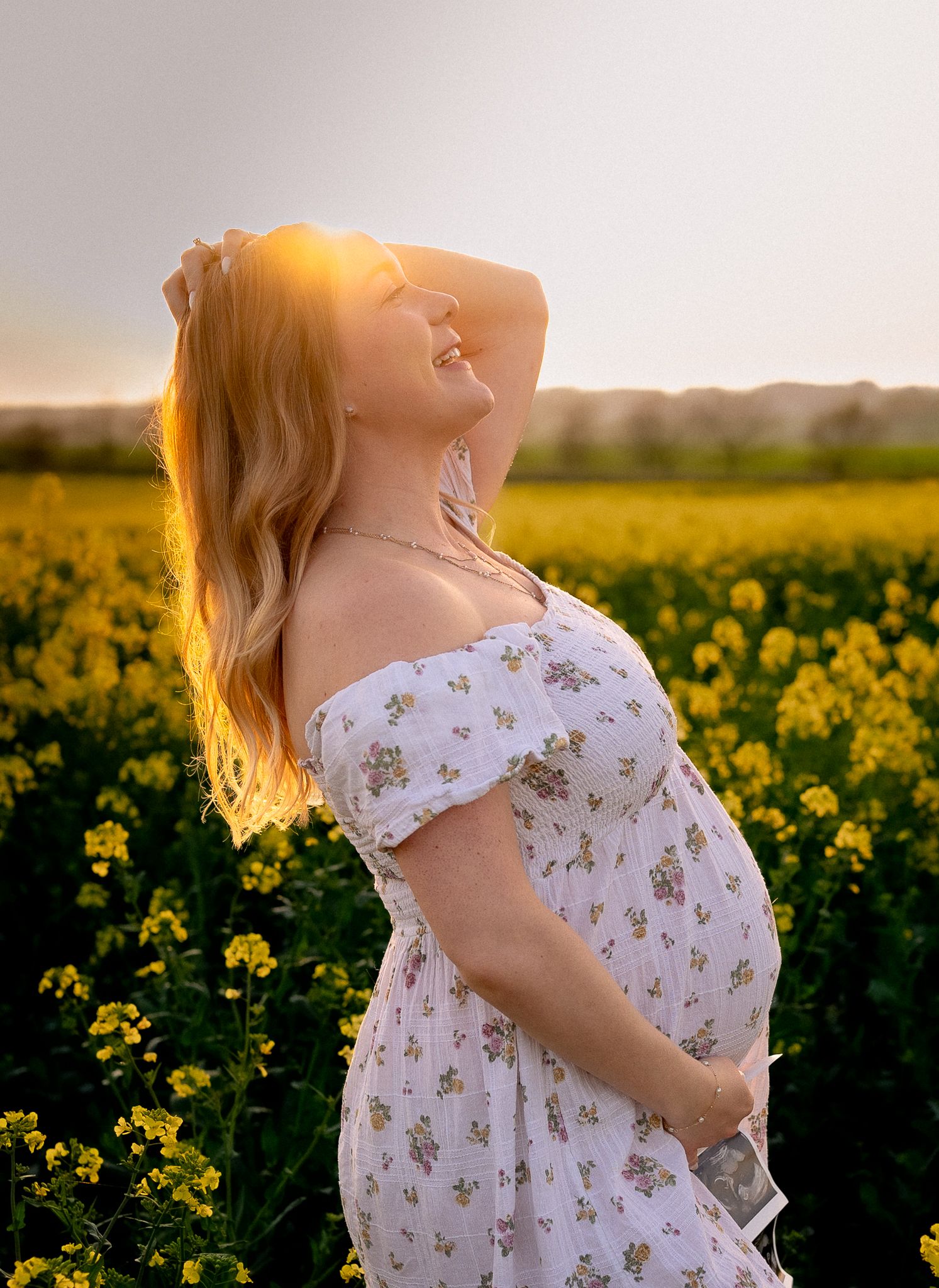 Pregnant woman in floral dress standing in yellow flower field at golden sunset, smiling with hand in hair.