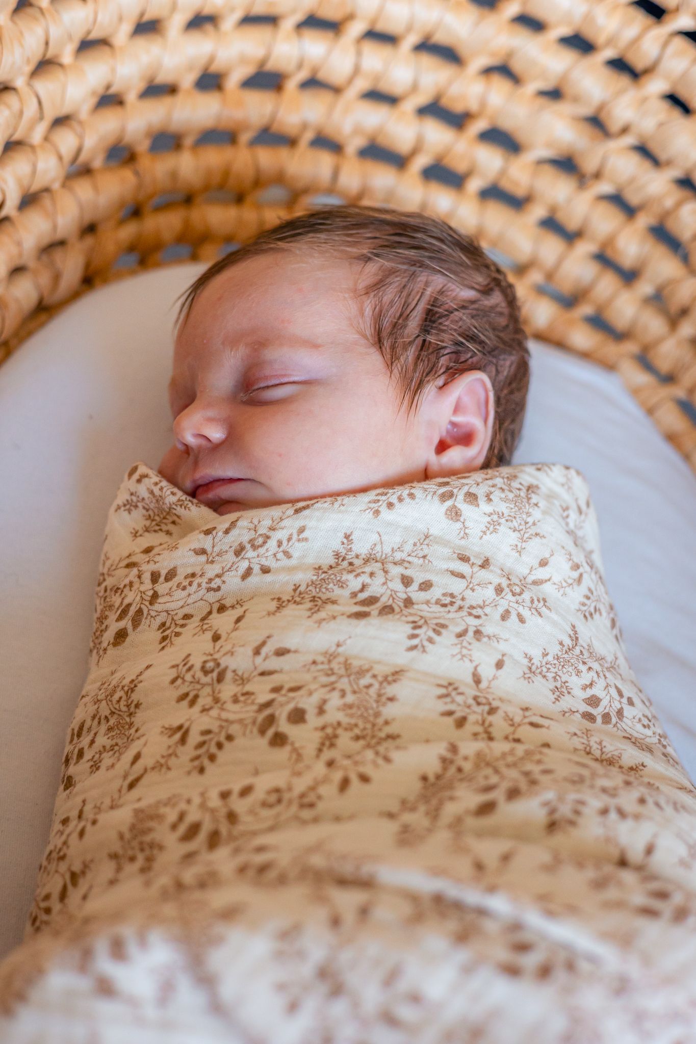 Relaxes, sleeping newborn wrapped in floral swaddle on woven basket background. Castle Combe
