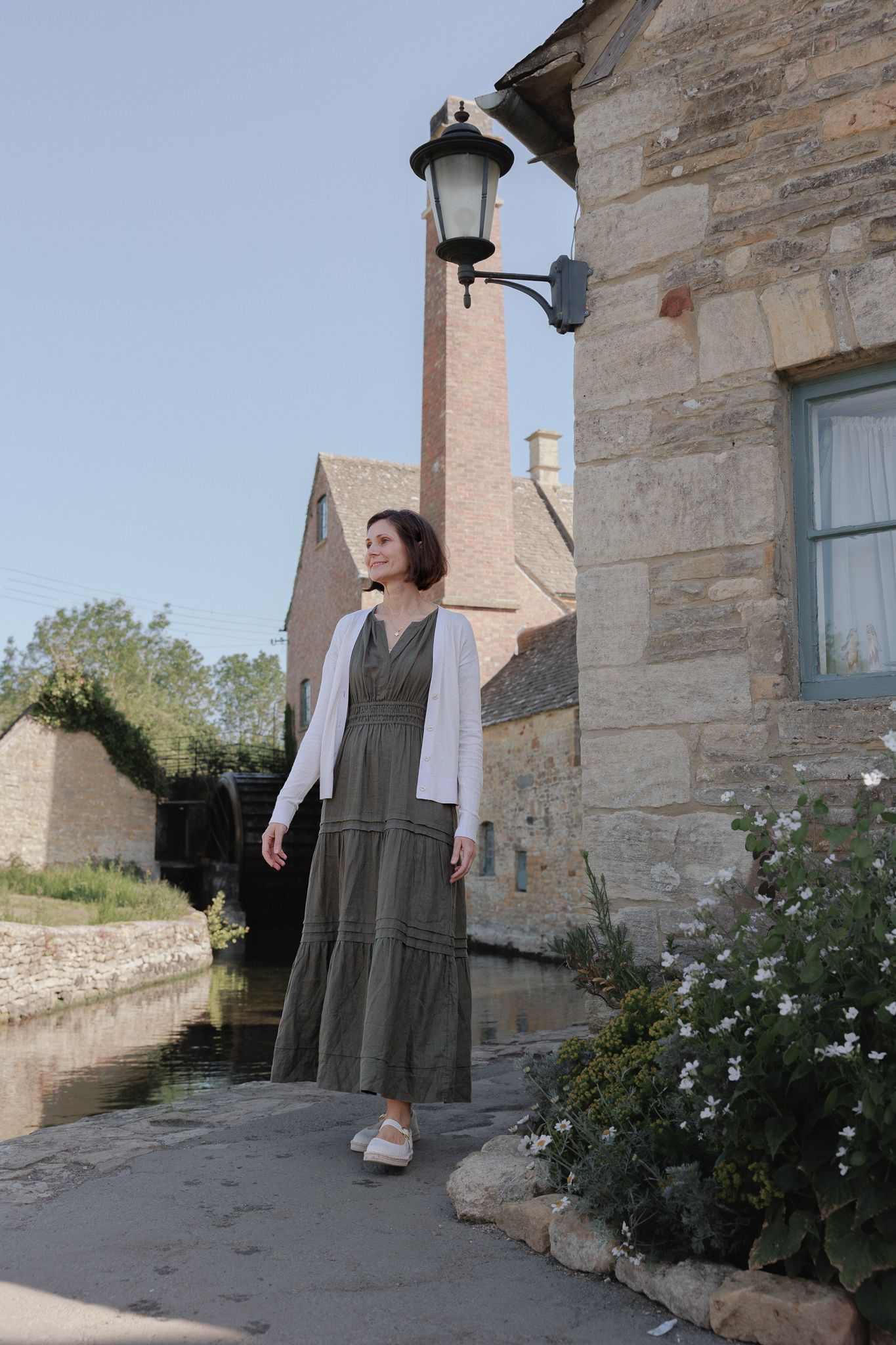 Woman in olive dress and white cardigan standing by a stone building and canal in a quaint English village.