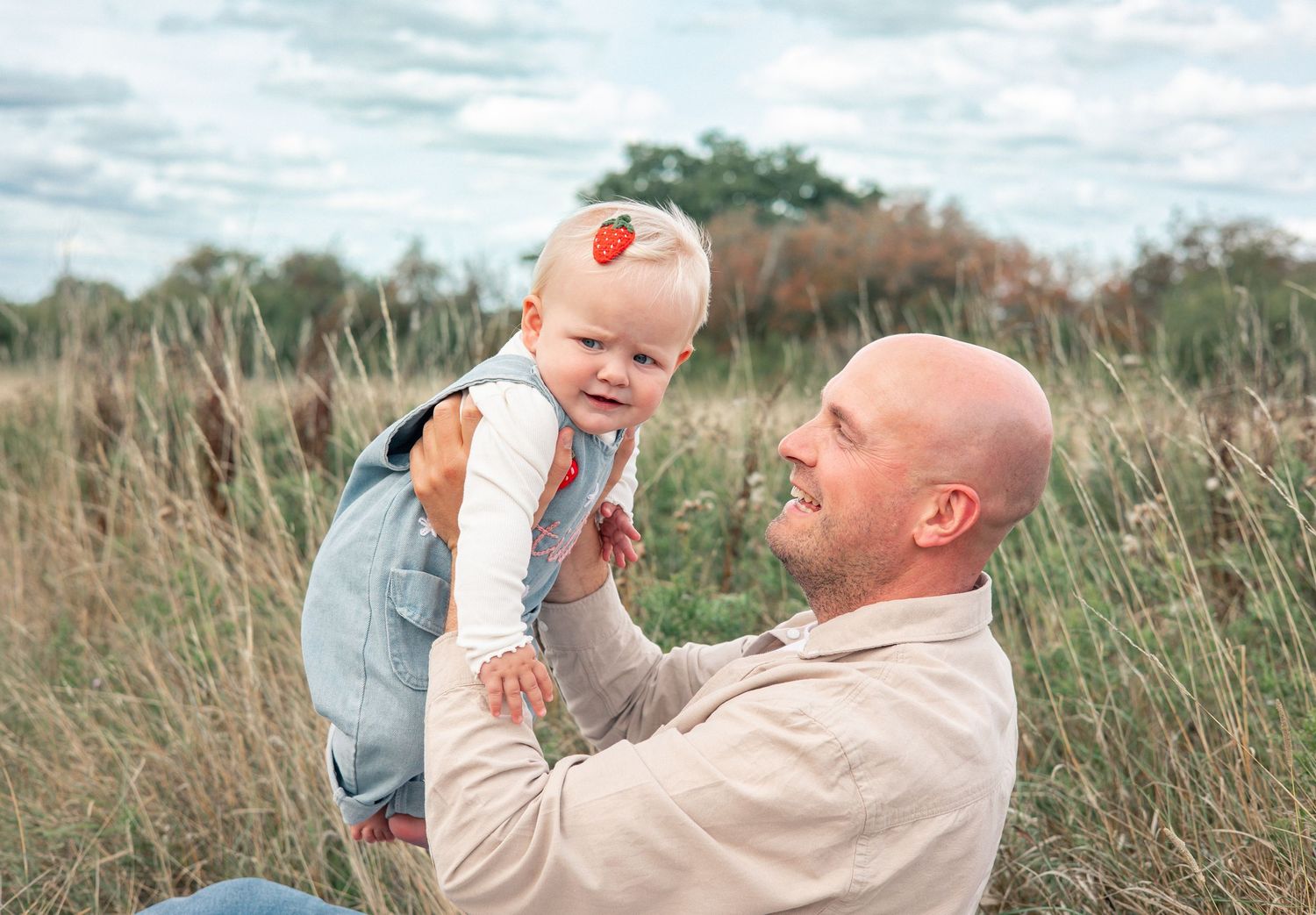 Bald man holding a smiling baby girl in a field of tall grass under a cloudy sky.