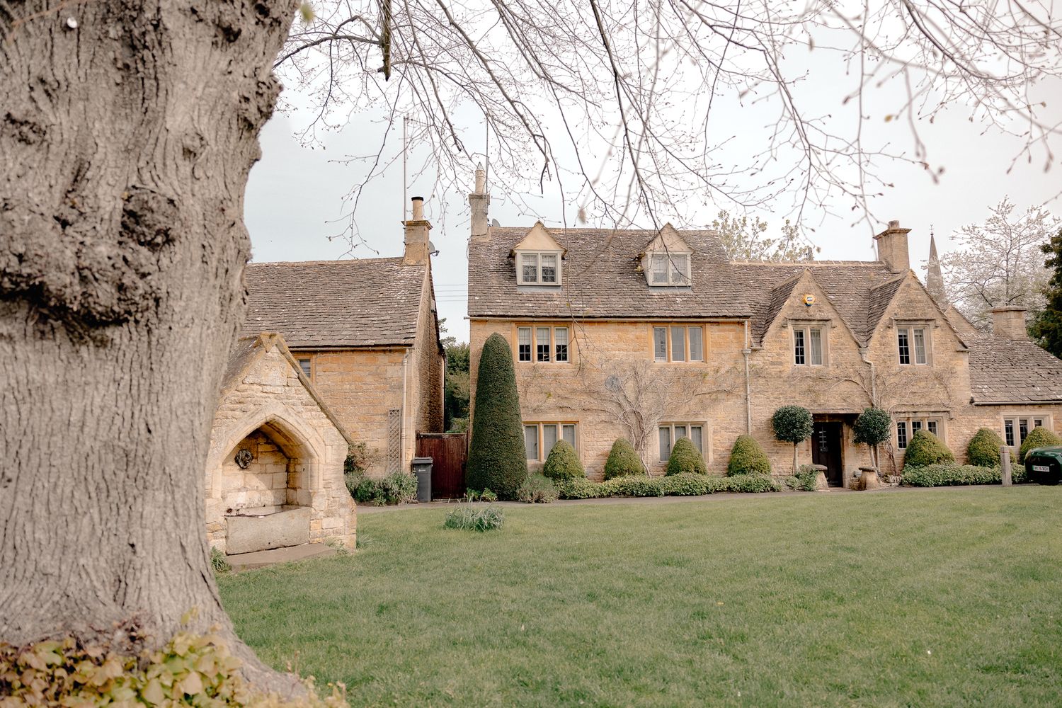 Charming Cotswold stone manor house with manicured lawn and mature tree in foreground in Lower Slaughter, Cotswolds, UK.