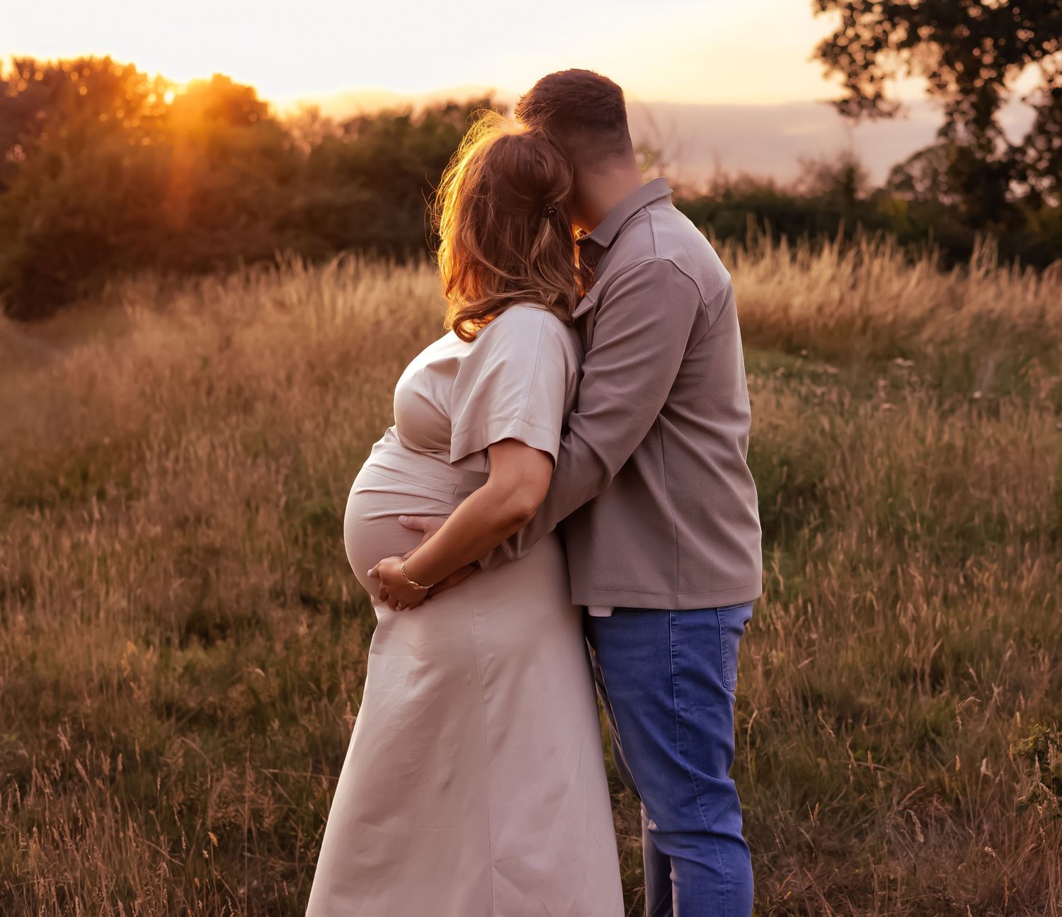 Couple sharing a tender kiss in a golden field at sunset, with pregnant woman cradling her baby bump in Cirencester.