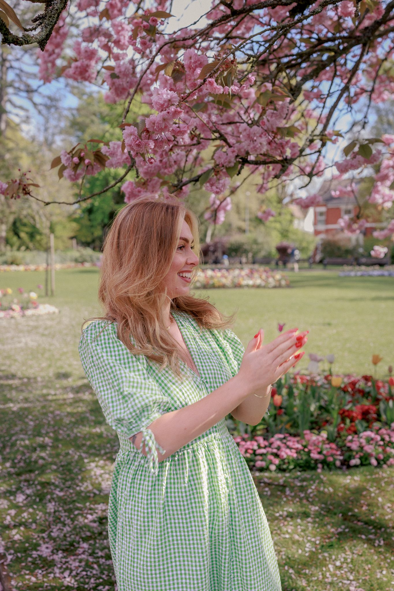 Woman in green gingham dress smiling under pink cherry blossom tree in a colourful spring park in Royal Wootton Bassett.