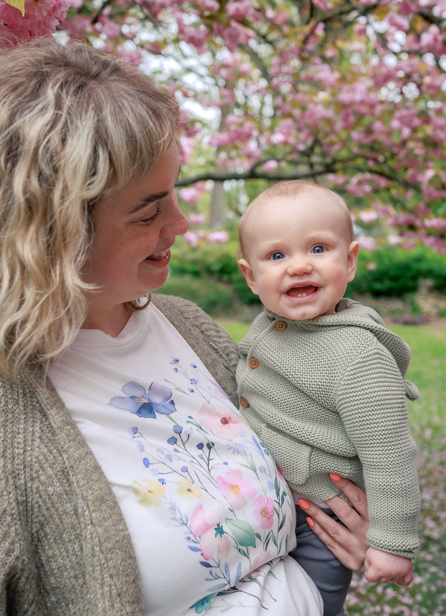 A smiling baby in a green knit hoodie held by a blonde woman in a floral shirt near pink cherry blossom trees in Painswick.