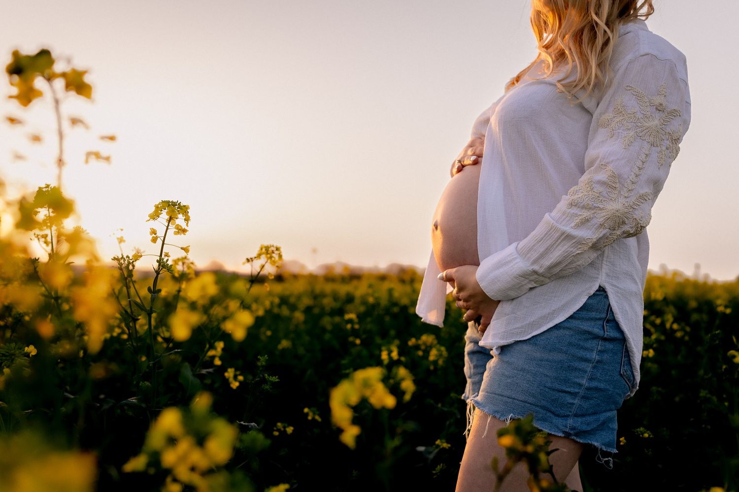 Pregnant woman in white shirt and denim skirt cradling baby bump in blooming yellow field at sunset in Oxford.