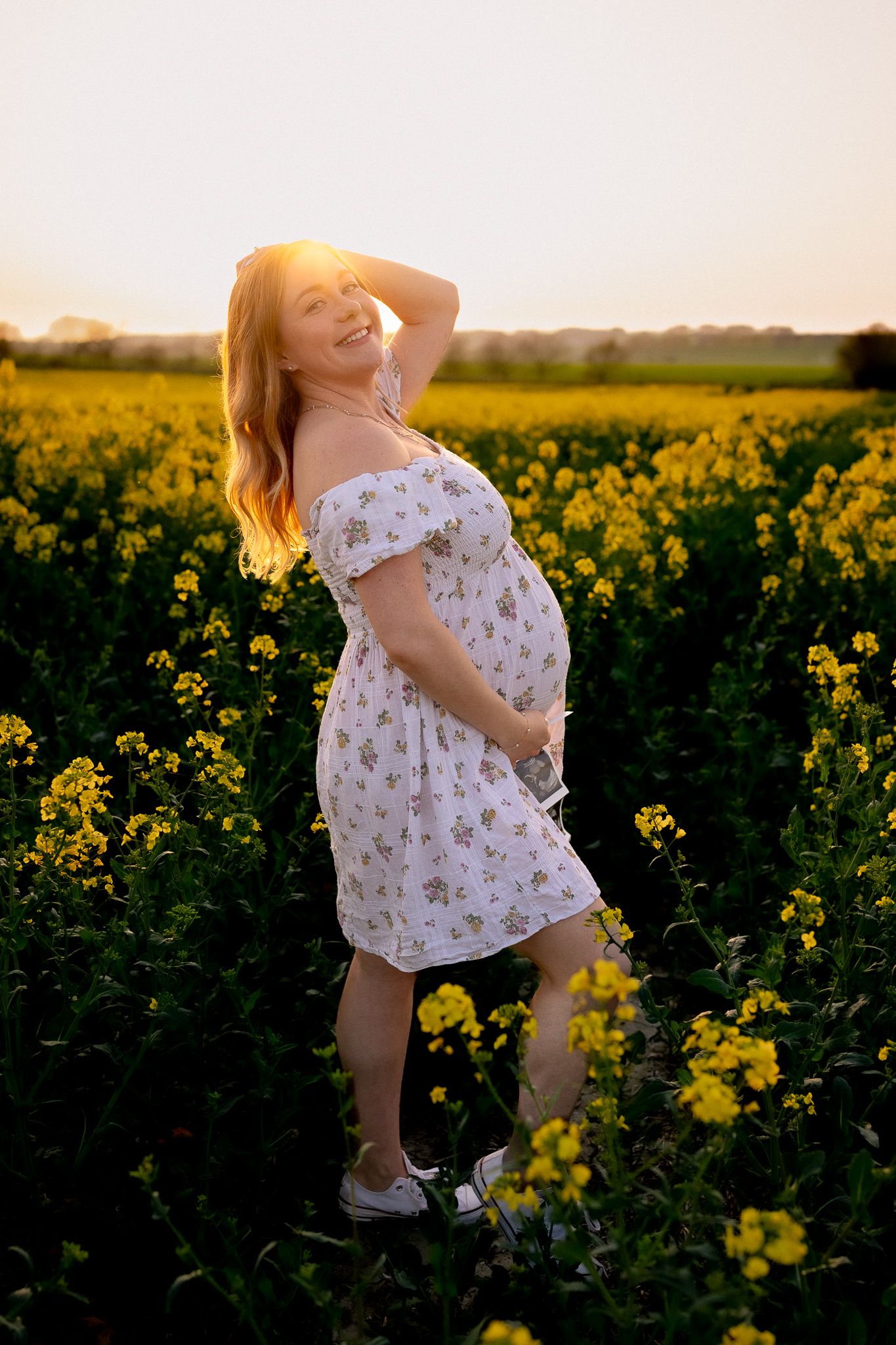 Pregnant woman in floral dress standing in yellow rapeseed field at golden sunset, smiling with hand in hair in Bath, UK.