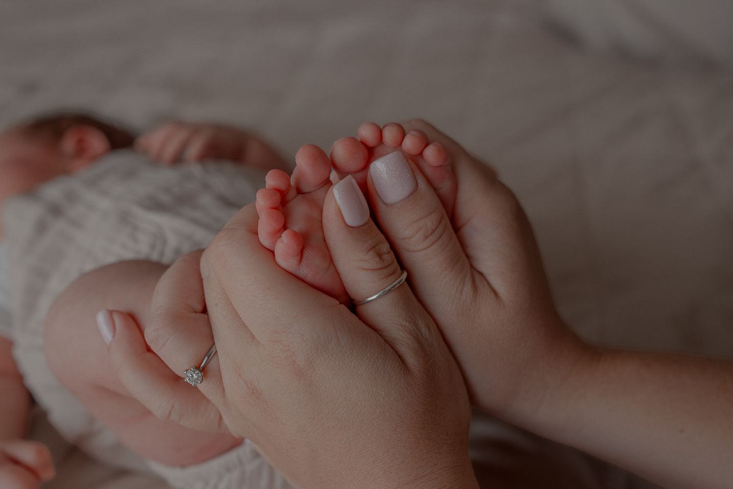 A mother gently holds her newborn baby's tiny feet in her hands, wearing a delicate ring, on a soft white blanket in Clifton, Bristol.