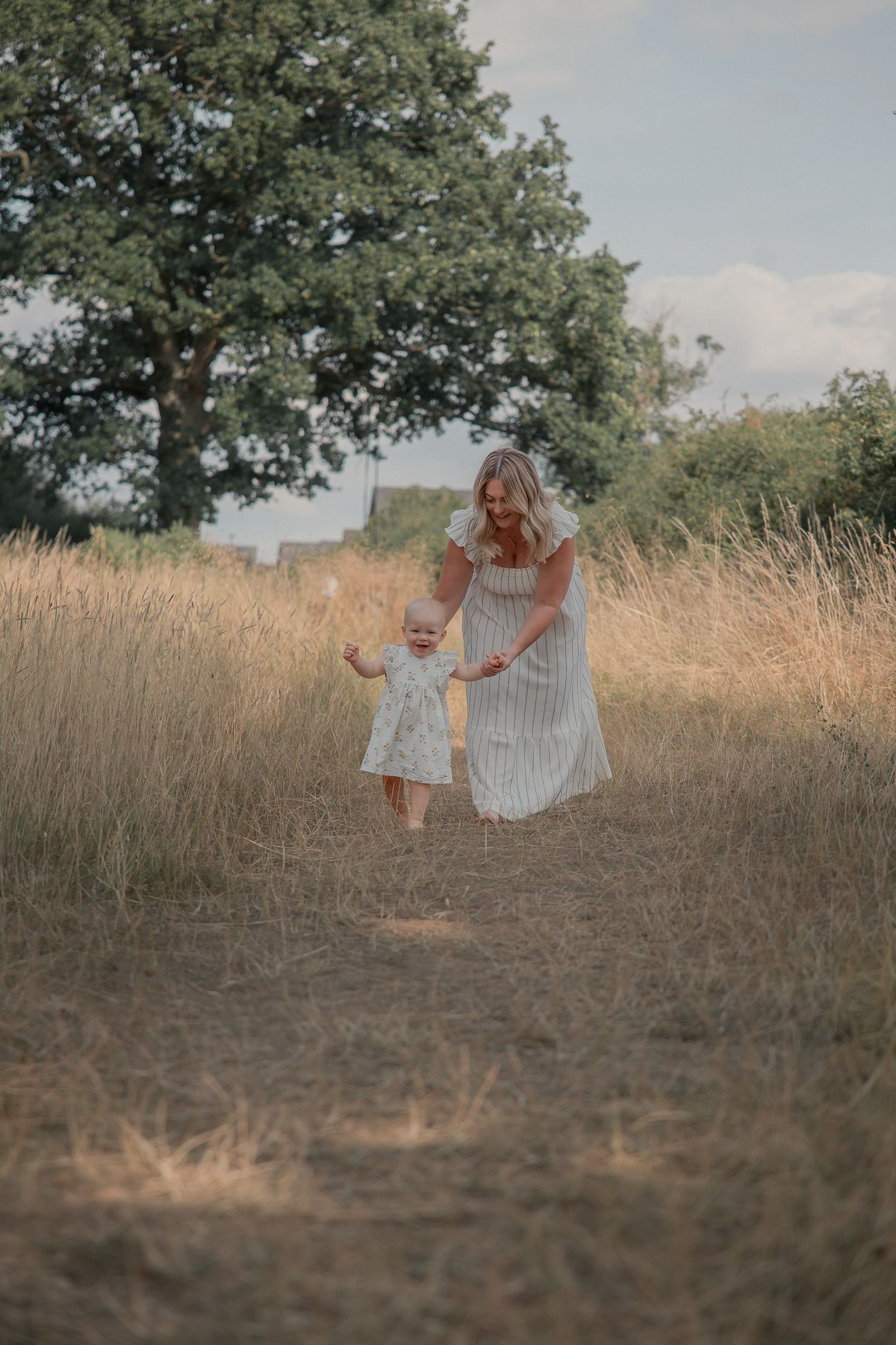 A woman in a striped dress helps a toddler walk along a golden grass field path beneath a large green tree.