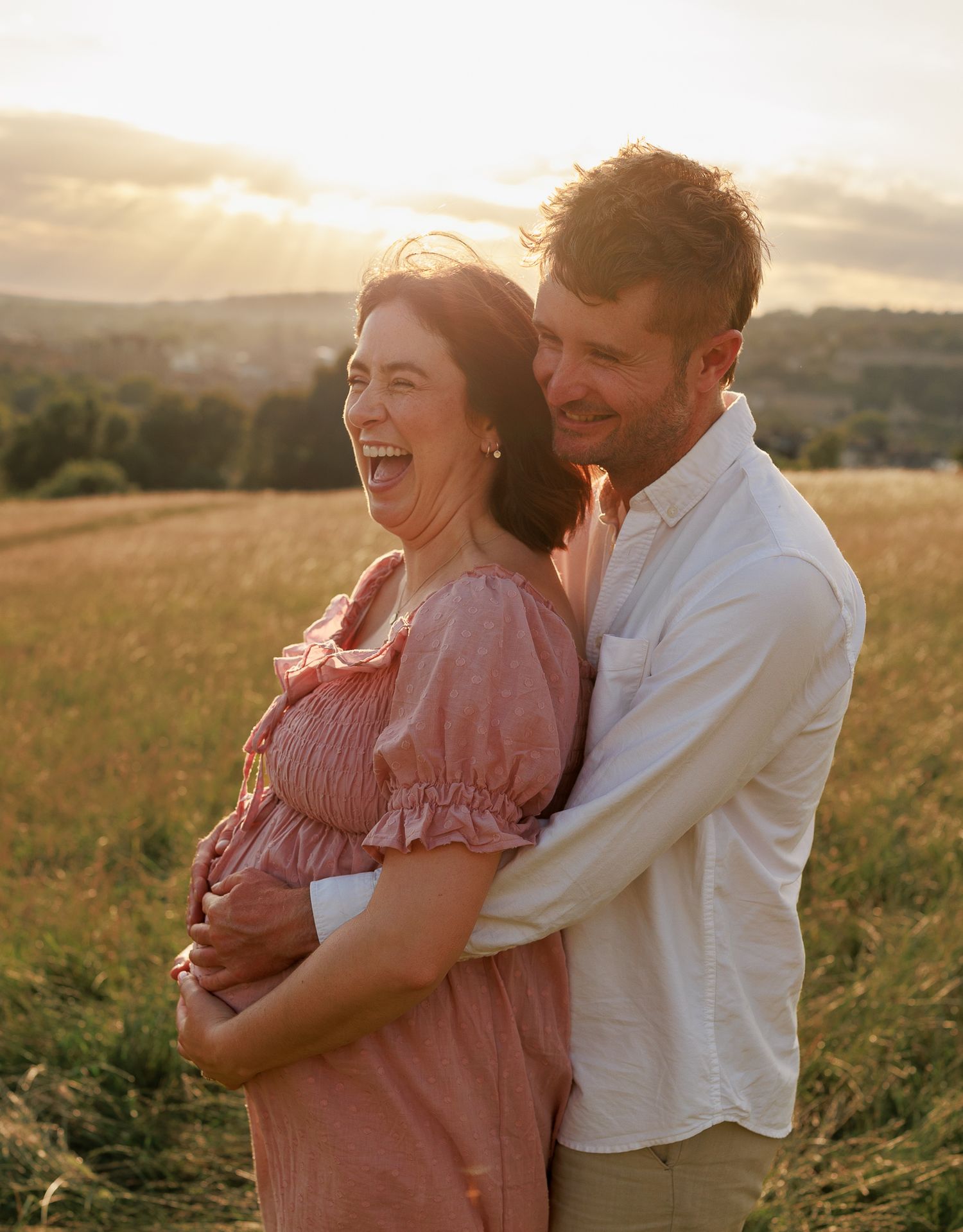 Laughing pregnant woman in pink dress embraced by man at golden sunset in green field for a Maternity Photoshoot in Wiltshire, UK.