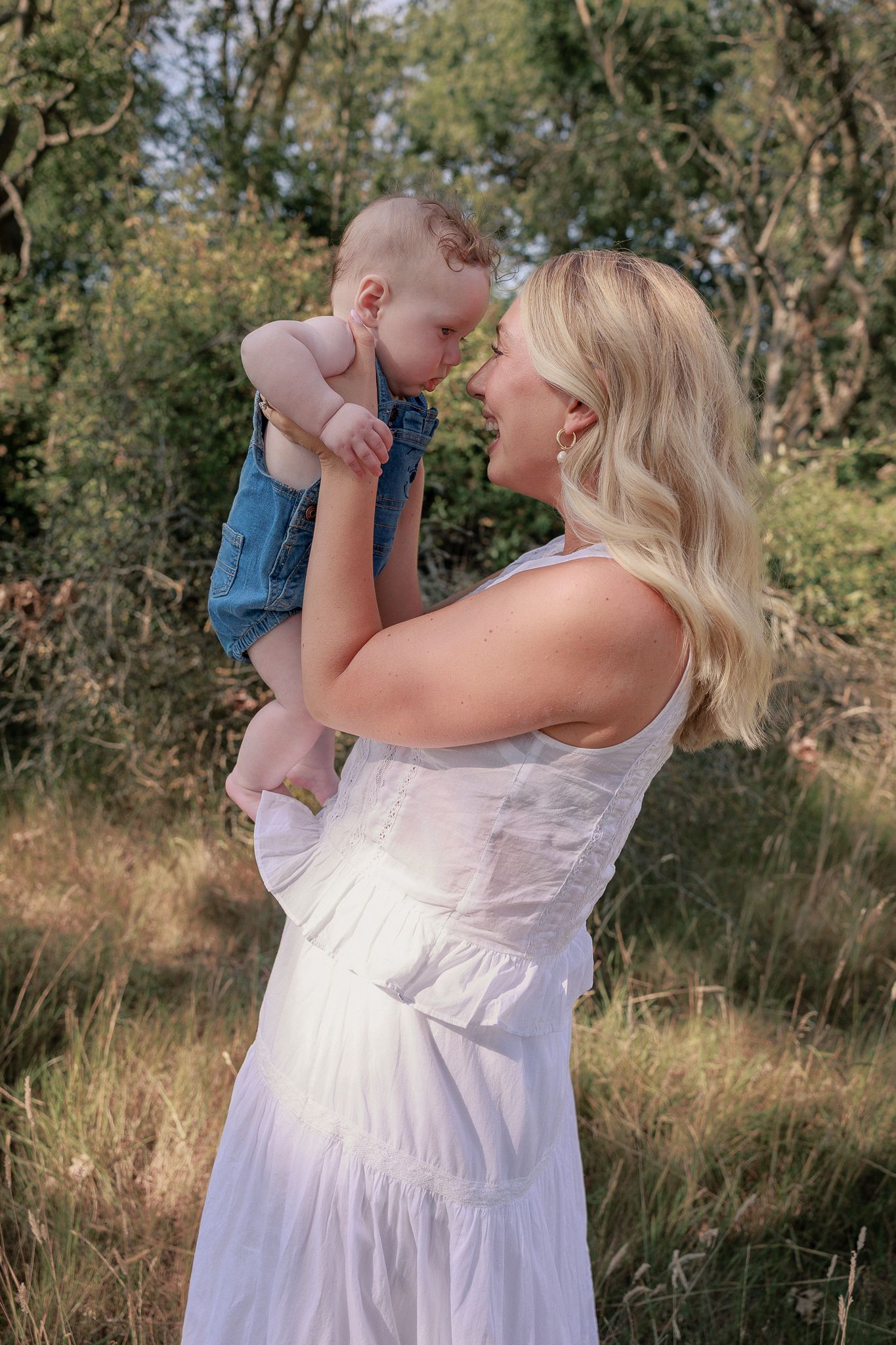 Blonde woman in white dress holding baby in denim overalls nose-to-nose in sunny outdoor setting in Stow, Cotswolds.