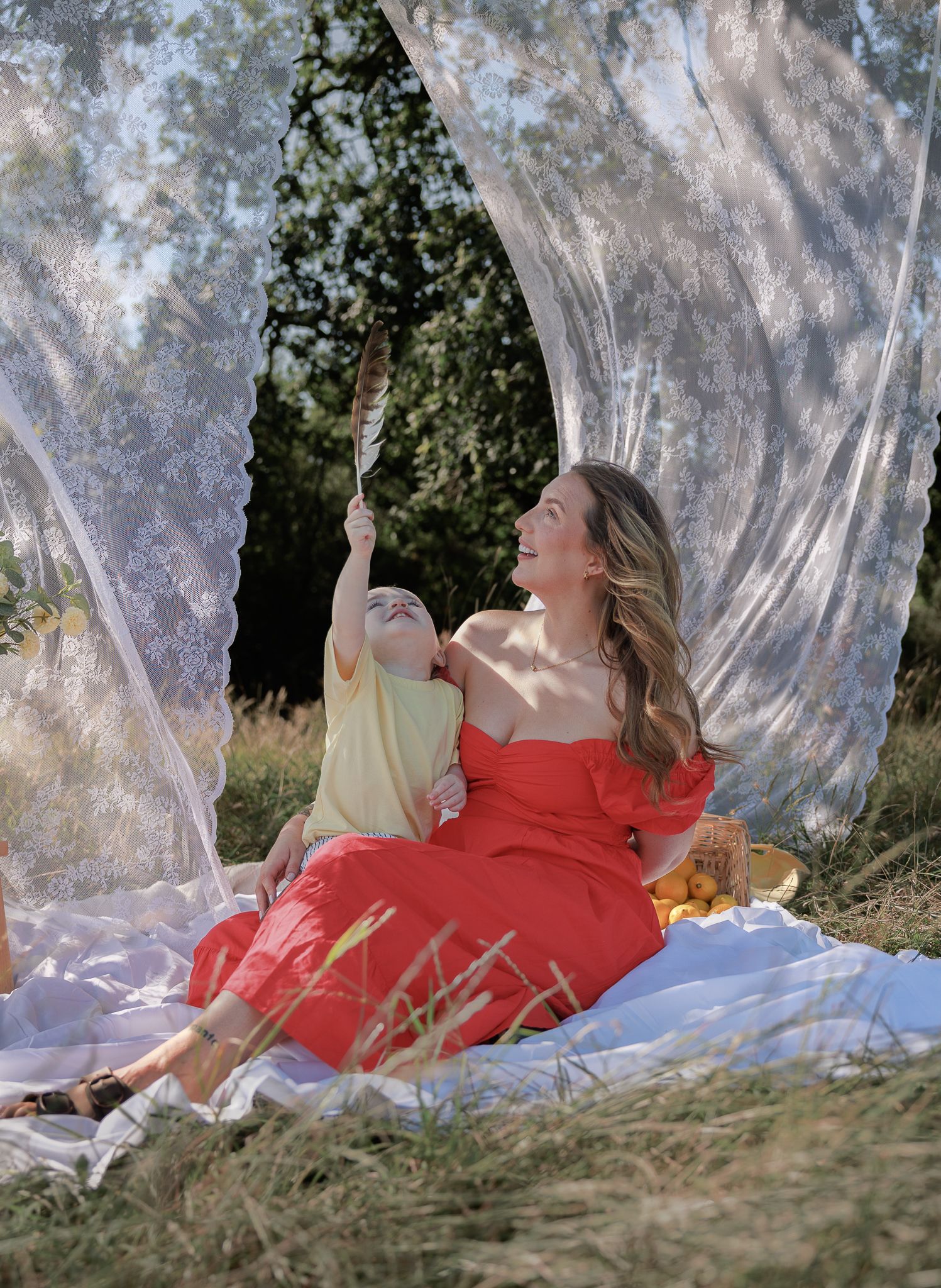 A woman in a red dress sits on a white blanket outdoors with a baby under flowing white curtains in Fairford, Cotswolds.