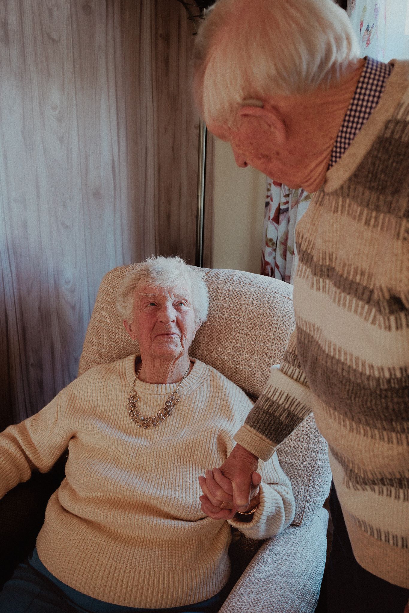 Two elderly people holding hands indoors, one seated in armchair looking up lovingly, sharing a tender moment in Upper Slaughter.
