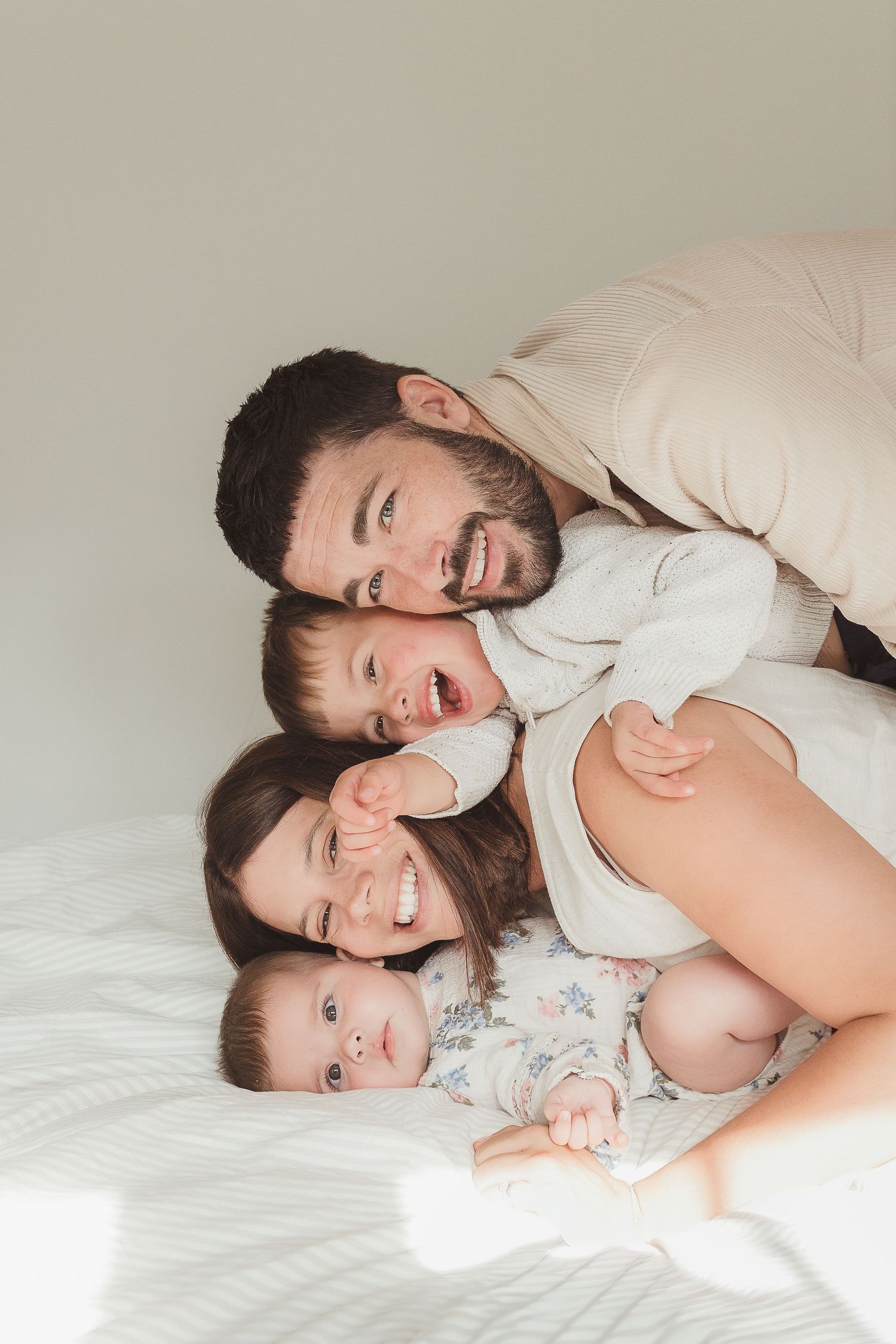 A happy family of four lying together on a white bed smiling in Malmesbury.