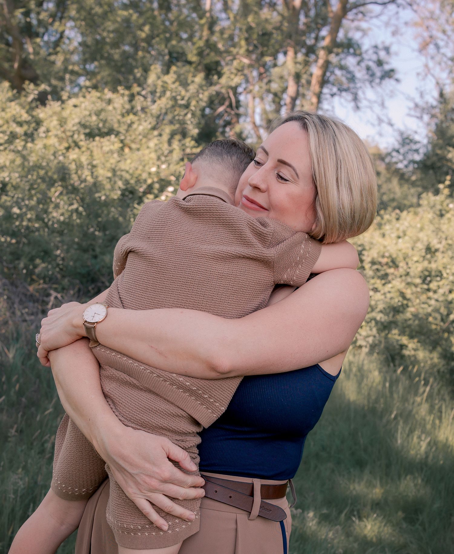 A woman with blonde hair embraces a person in military uniform outdoors in a sunlit field with trees in the background.