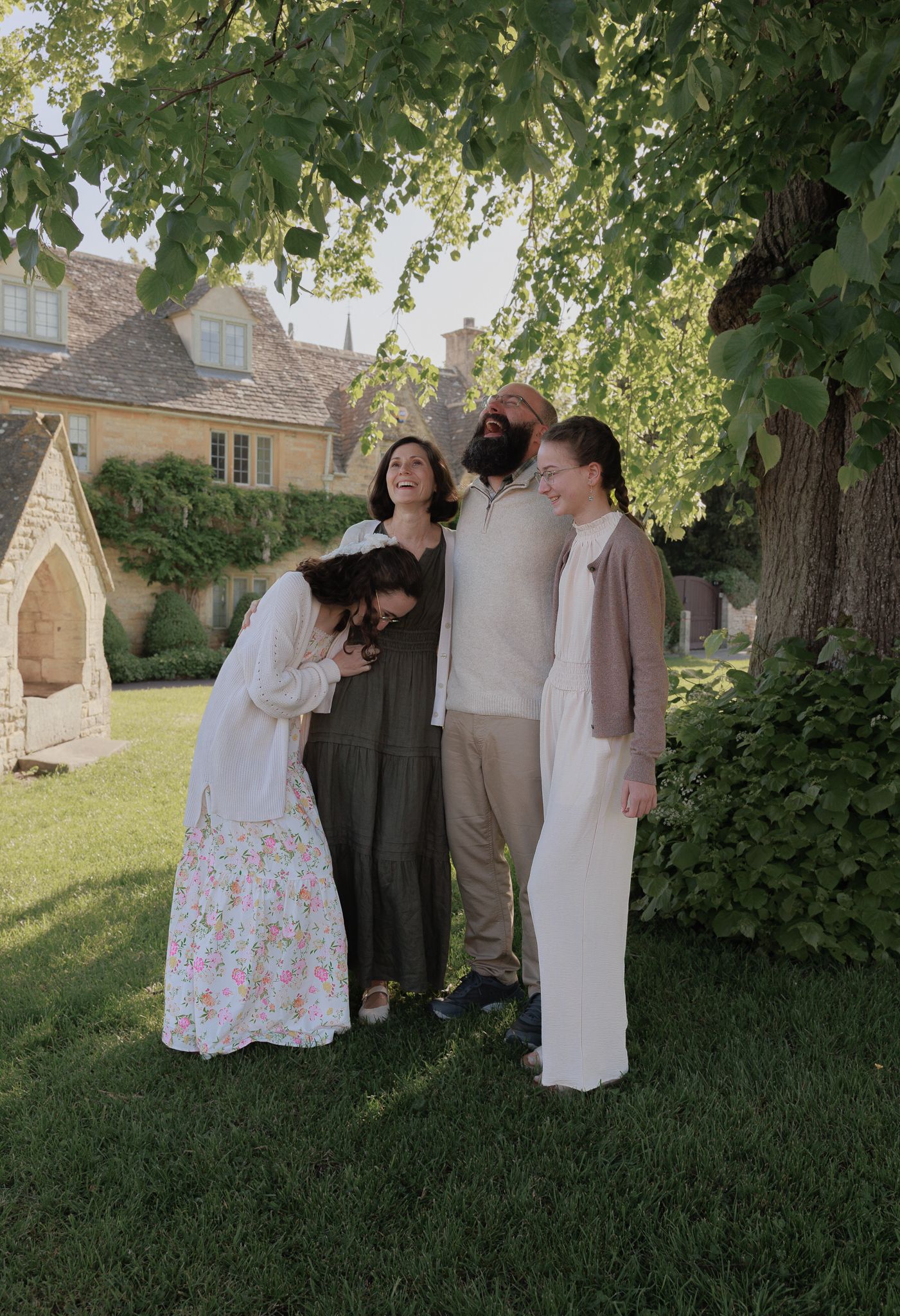 Family of four laughing together outdoors on green lawn beside stone cottage under leafy tree in warm summer light near The Slaughters.