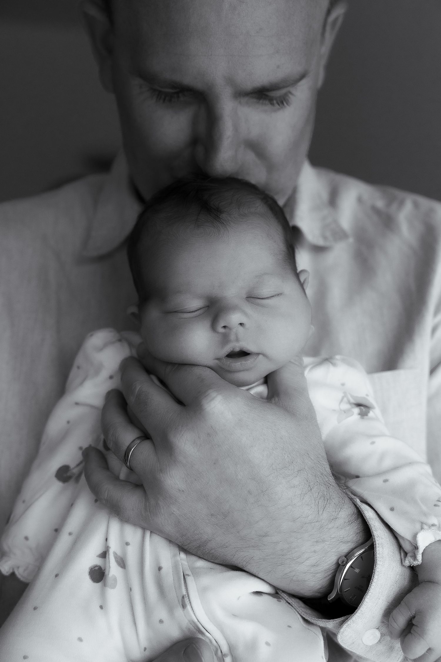 Black and white portrait of a father in a button-up shirt gently kissing the head of his sleeping newborn baby in a polka dot outfit.