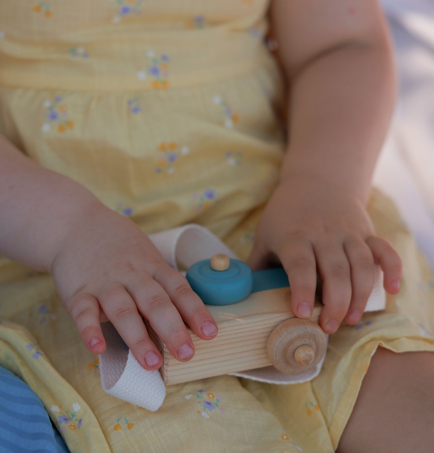 Baby in yellow floral dress playing with a wooden toy car, close-up of tiny hands holding colorful wooden toy in Bibury, Cotswolds.