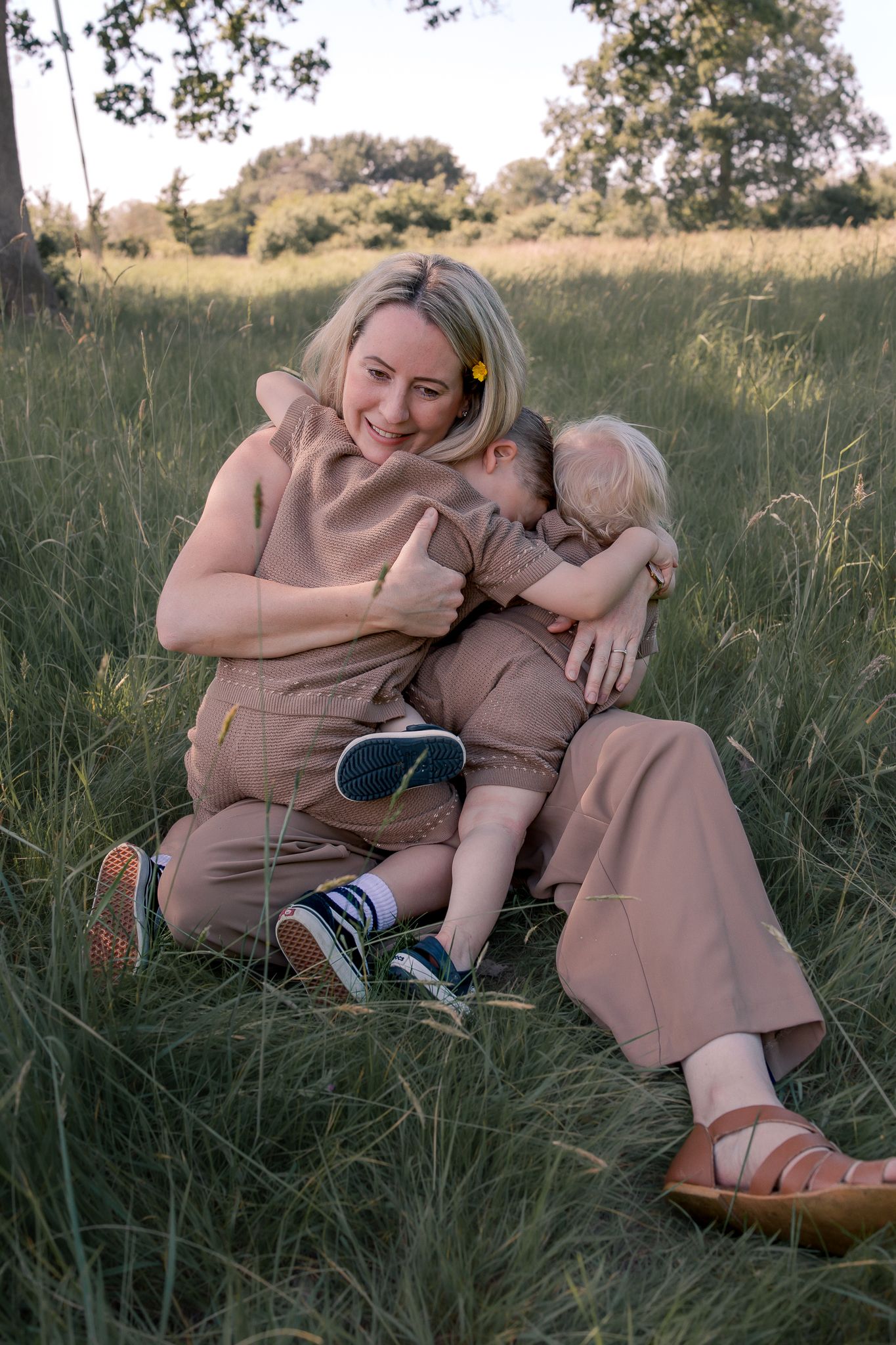 Mother and child sitting together in a grassy field during golden hour, wearing matching beige outfits and sneakers.